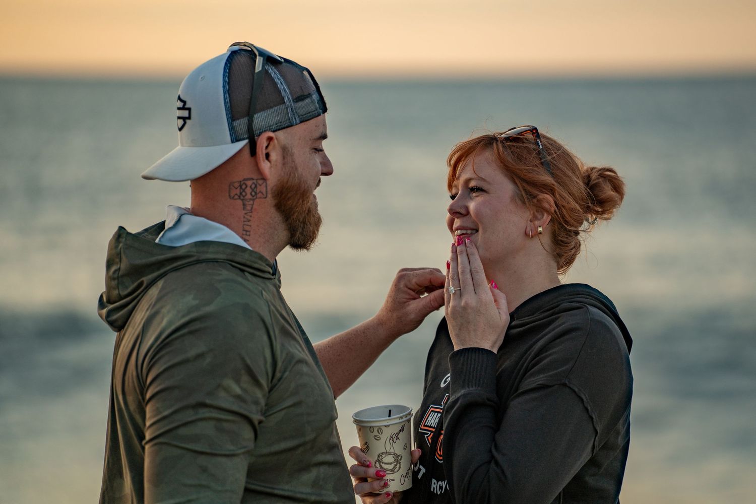 Smiling woman with red hair covers her mouth as a bearded man in a cap and green hoodie reaches toward her on a beach at sunset.