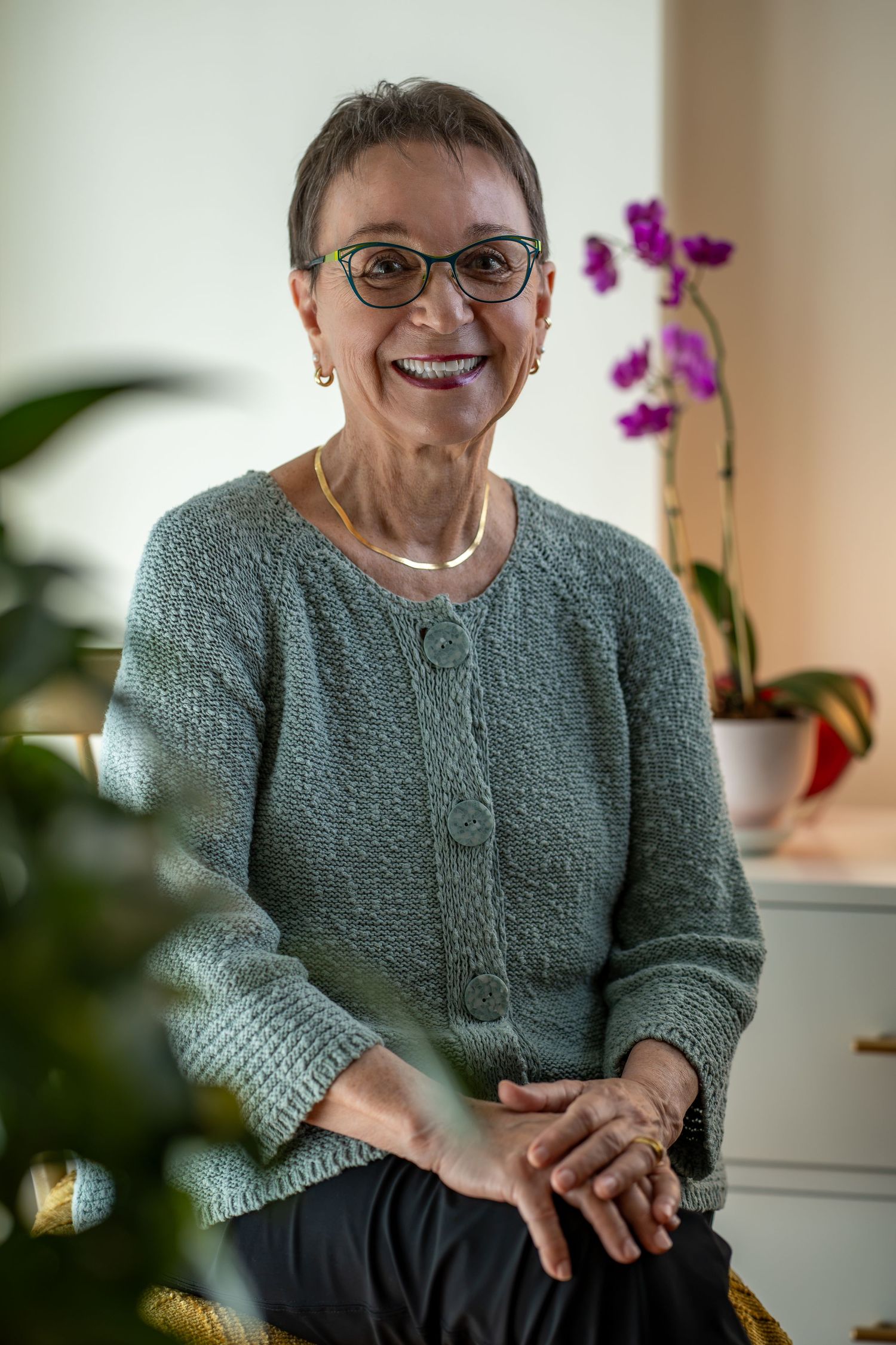 Smiling senior woman with short hair and glasses, seated indoors with orchids in background, author headshot, Boston Headshot Photographer