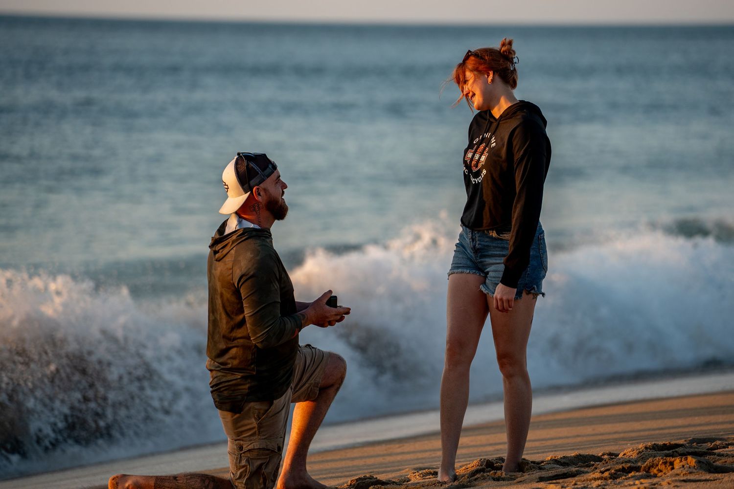 Man in a backwards cap kneels on one knee holding a ring box as a smiling woman looks on during a beach marriage proposal at sunset.