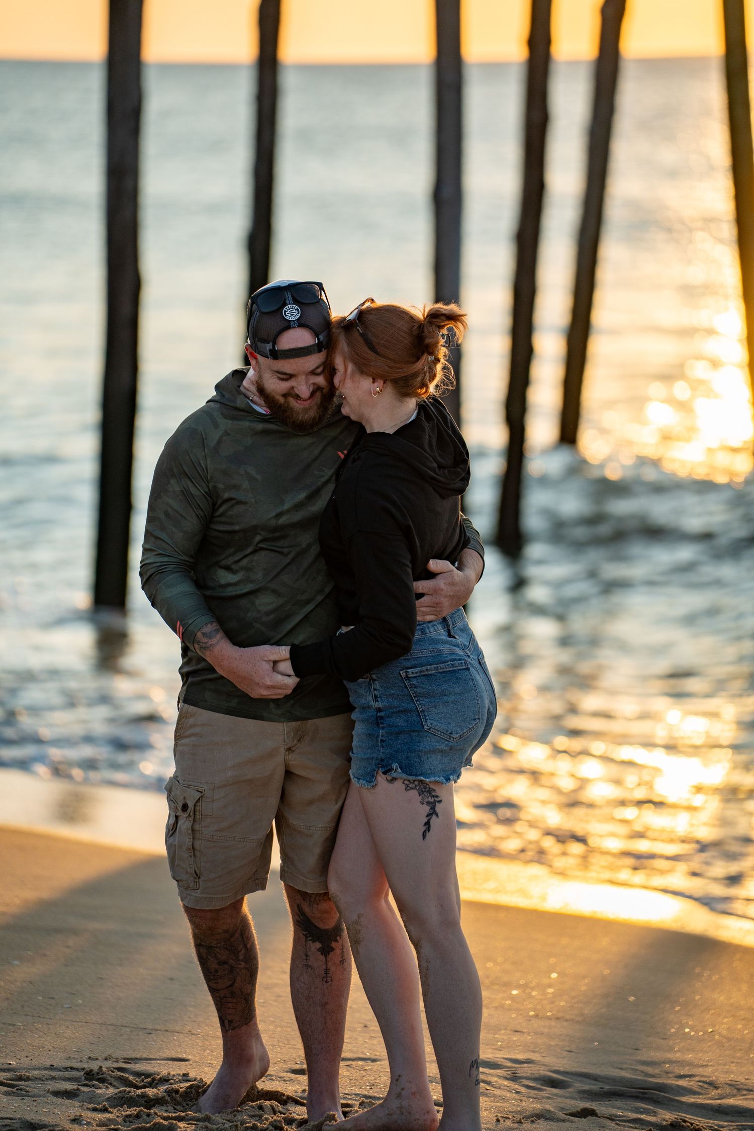 Smiling couple embracing on a sandy beach at sunset, standing near wooden pier pilings with golden light sparkling on the ocean water.