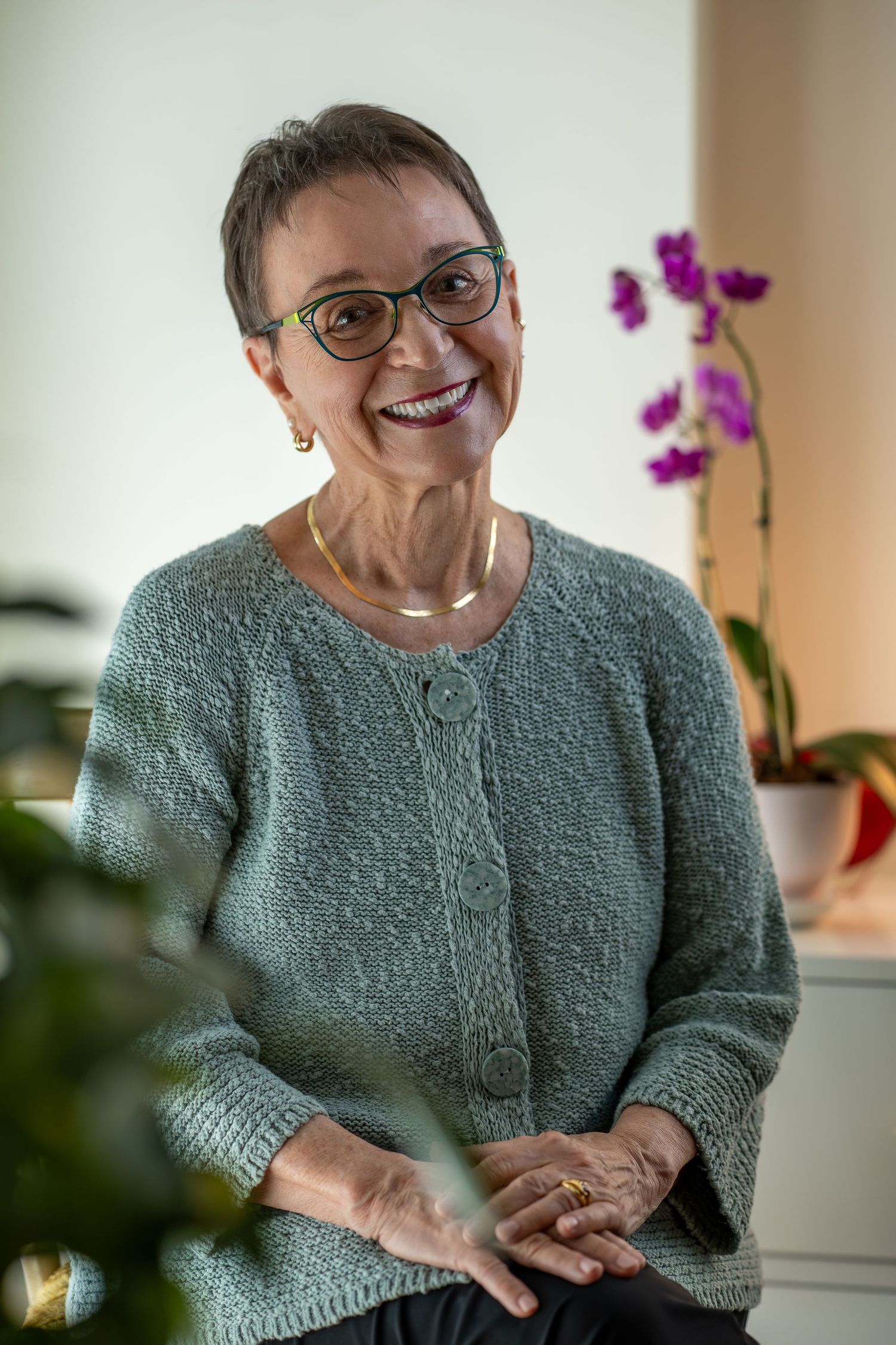 Older woman with short hair and green glasses smiling in teal cardigan near purple orchids.