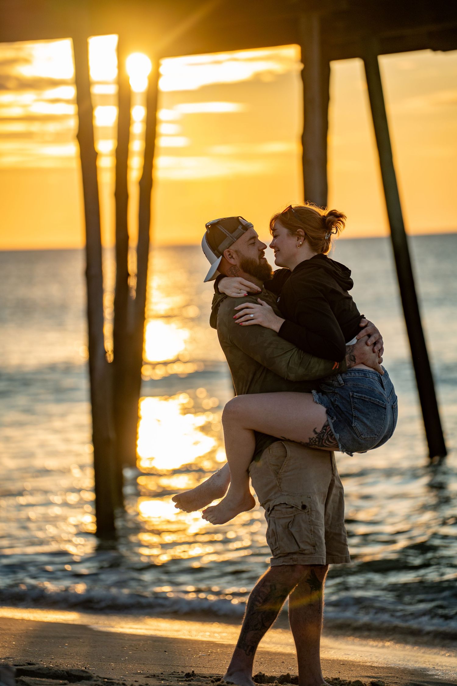 Couple embracing on a sandy beach at sunset as a man lifts a smiling woman beneath a wooden pier with golden light on the ocean.