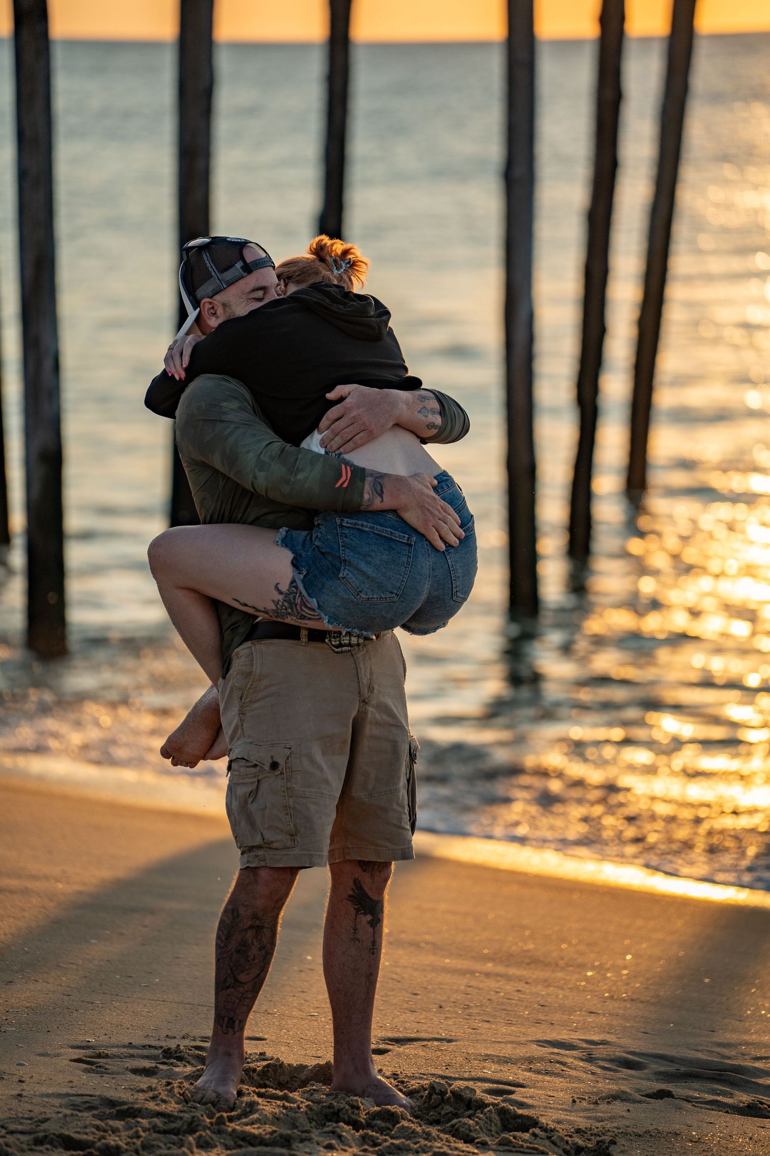 Tattooed man in cargo shorts lifts and hugs a woman on a sandy beach at sunset, with wooden pier pilings and golden light on the water.