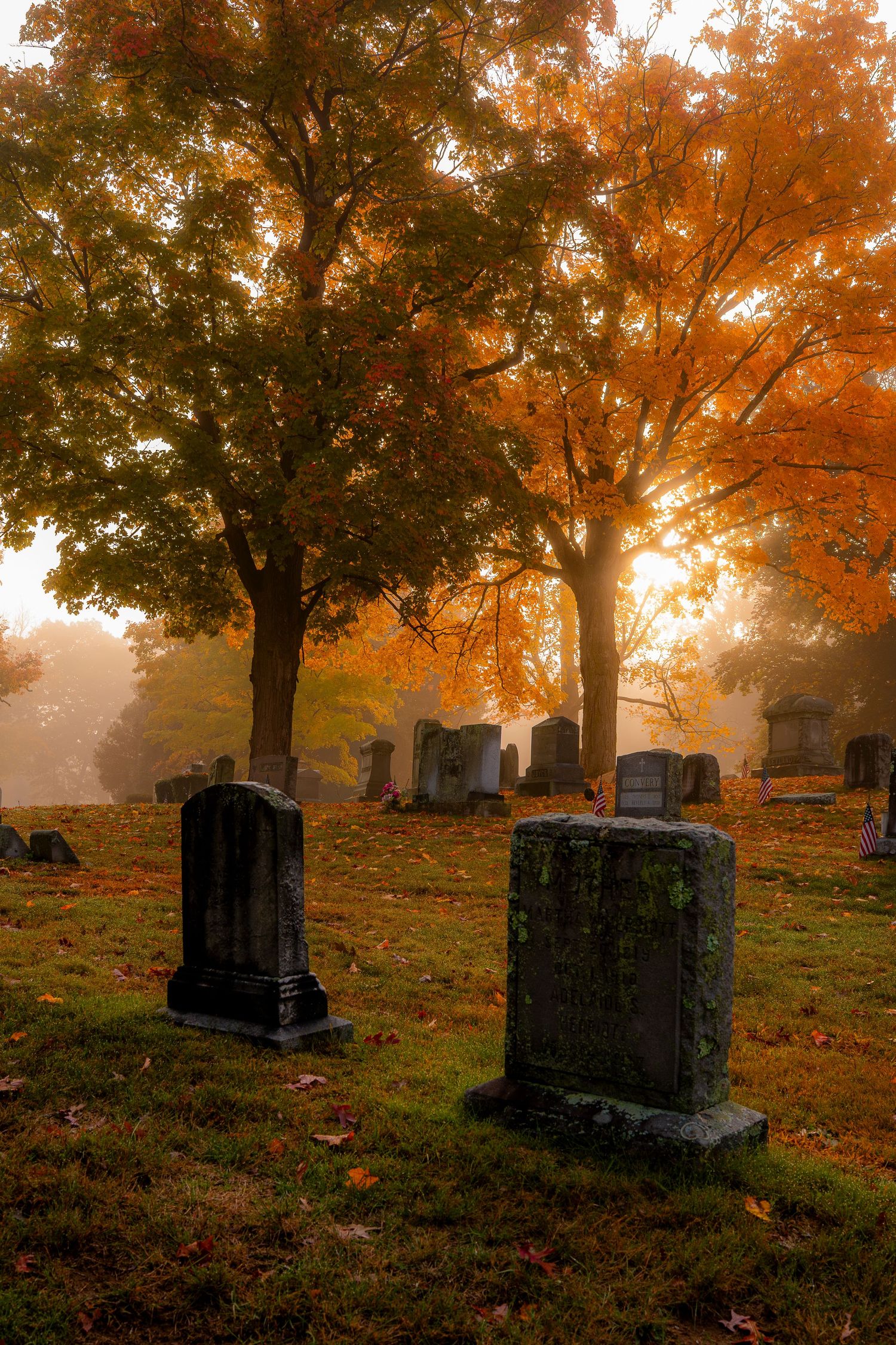 Misty autumn cemetery with moss-covered gravestones, vibrant orange foliage, and soft golden sunlight filtering through foggy trees.
