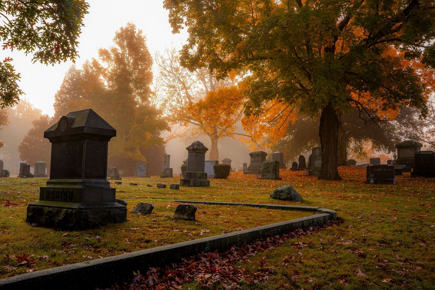 Misty autumn cemetery with old stone grave markers surrounded by colorful fall foliage trees in golden morning light.