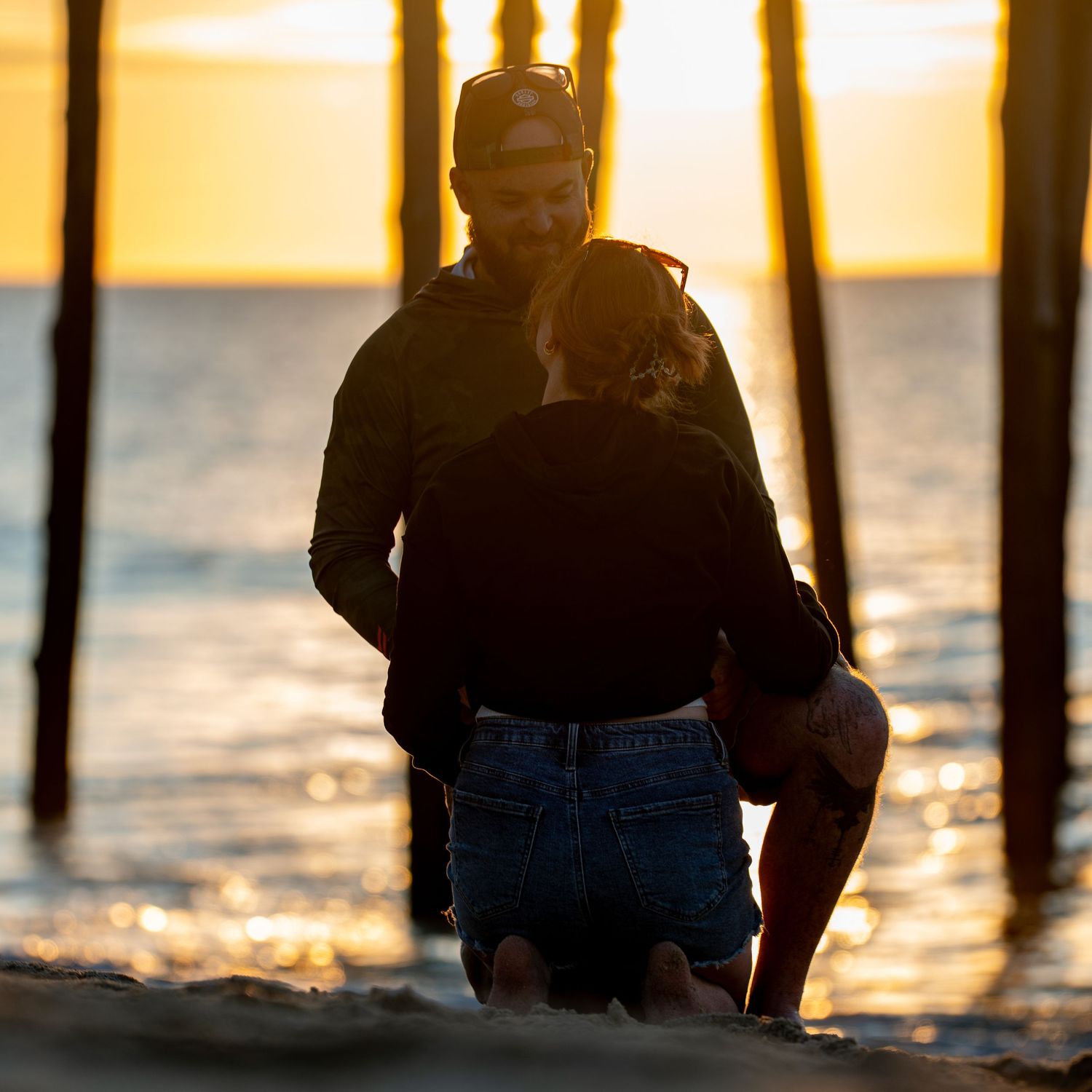 Silhouetted couple embracing on a sandy beach at golden sunset, with wooden pier pilings and sparkling ocean waves in the background.