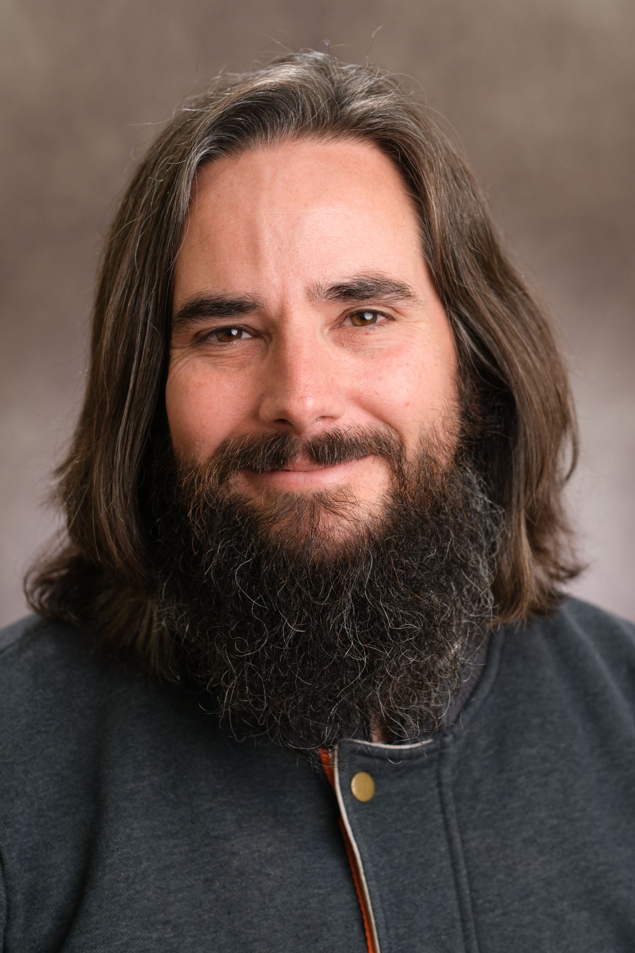 A bearded man with long brown hair smiles warmly in a professional headshot portrait, Tom Scholfield, Portrait & Headshot Photographer