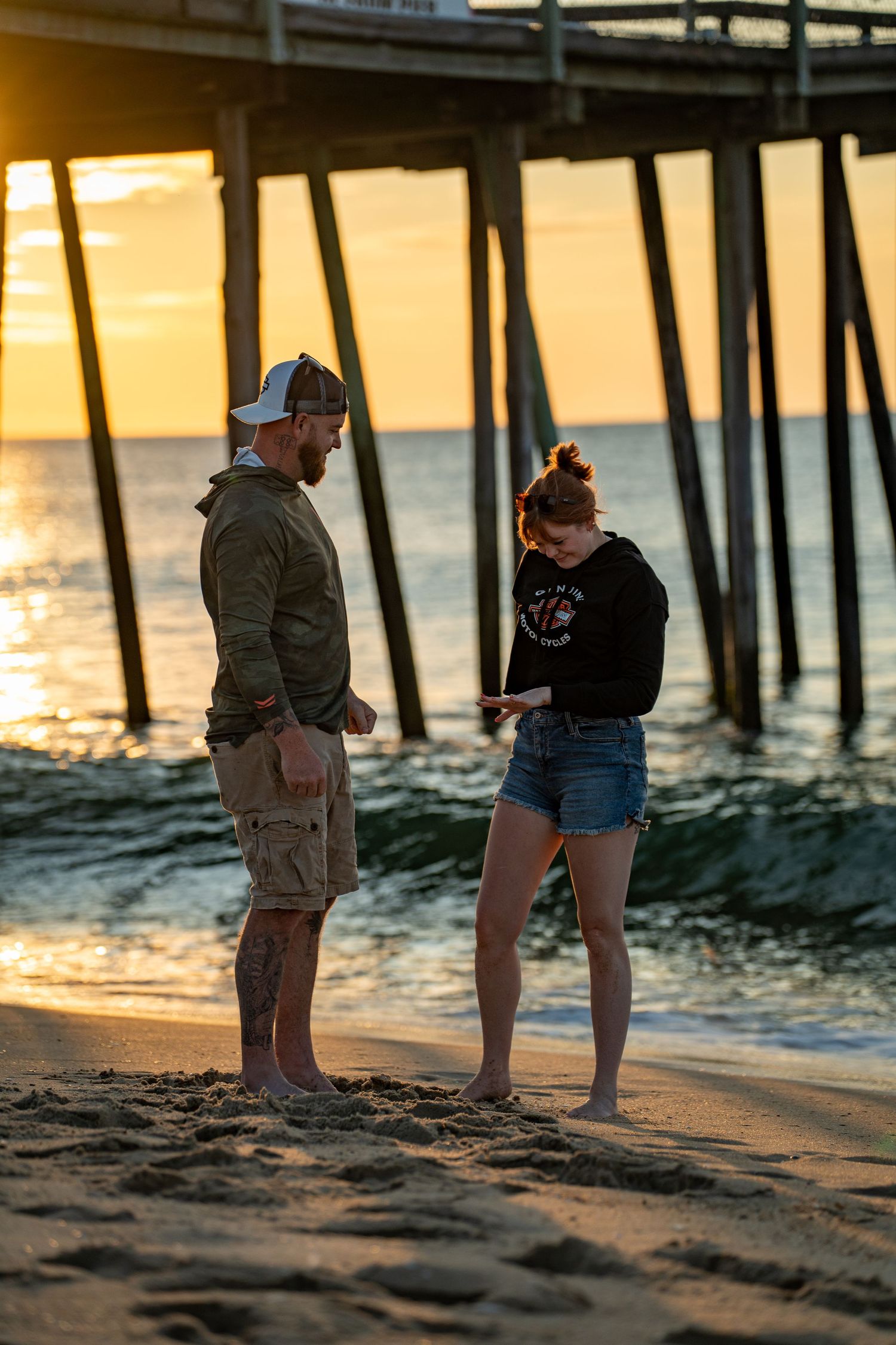 A bearded man in a cap and a woman in denim shorts stand barefoot on a sandy beach at sunset beside a wooden fishing pier.