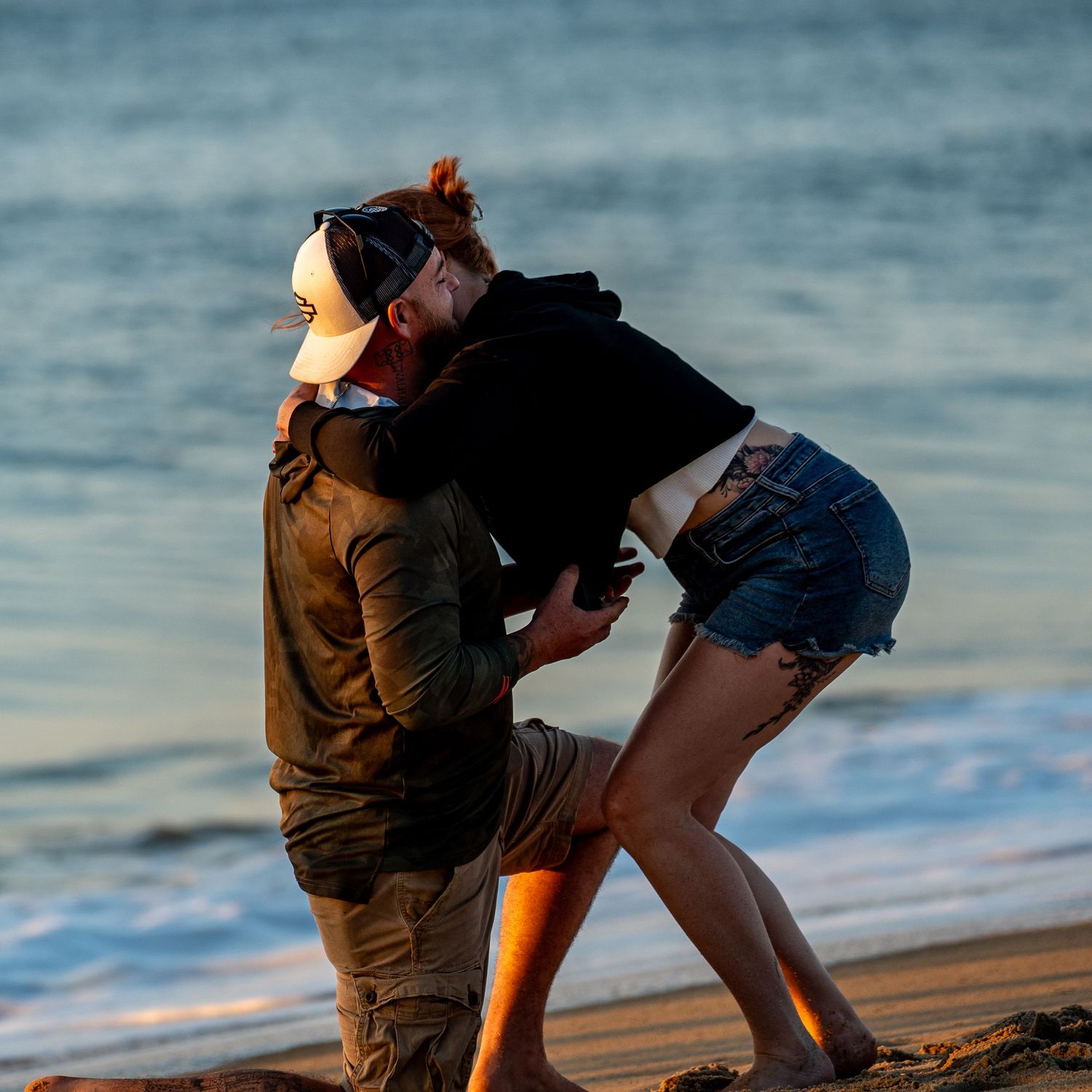A man in a trucker hat kneels on a sandy beach at sunset, embracing a woman in denim shorts as gentle ocean waves roll in behind them.