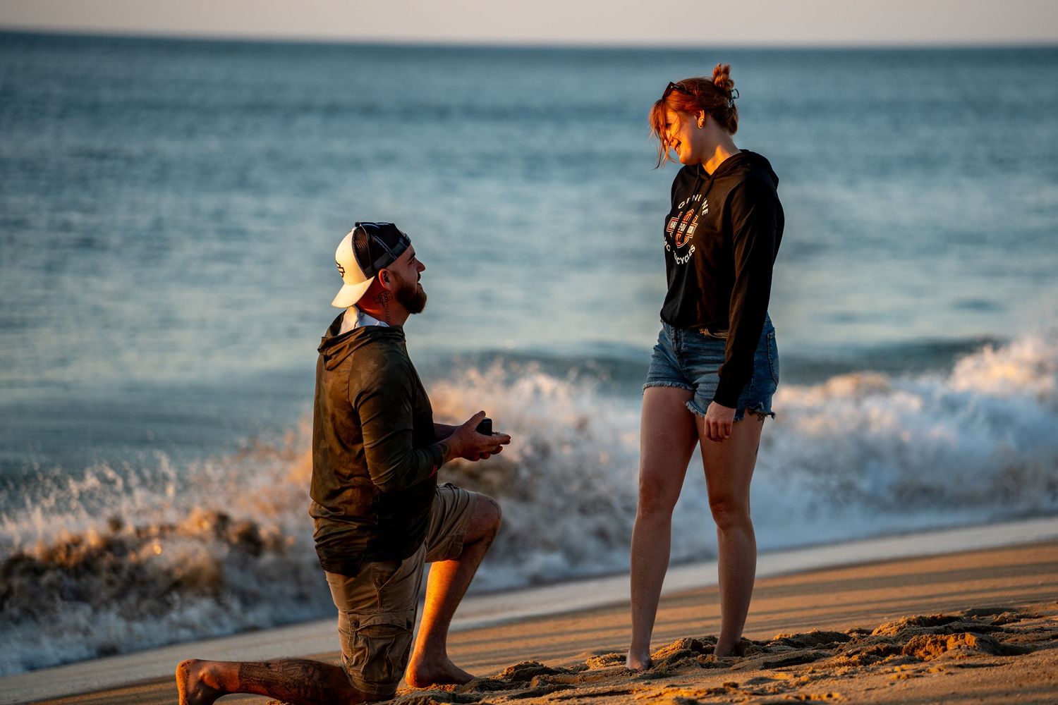 Man kneeling on one knee holding a ring box proposes to a smiling woman standing on a sunset beach with ocean waves behind them.