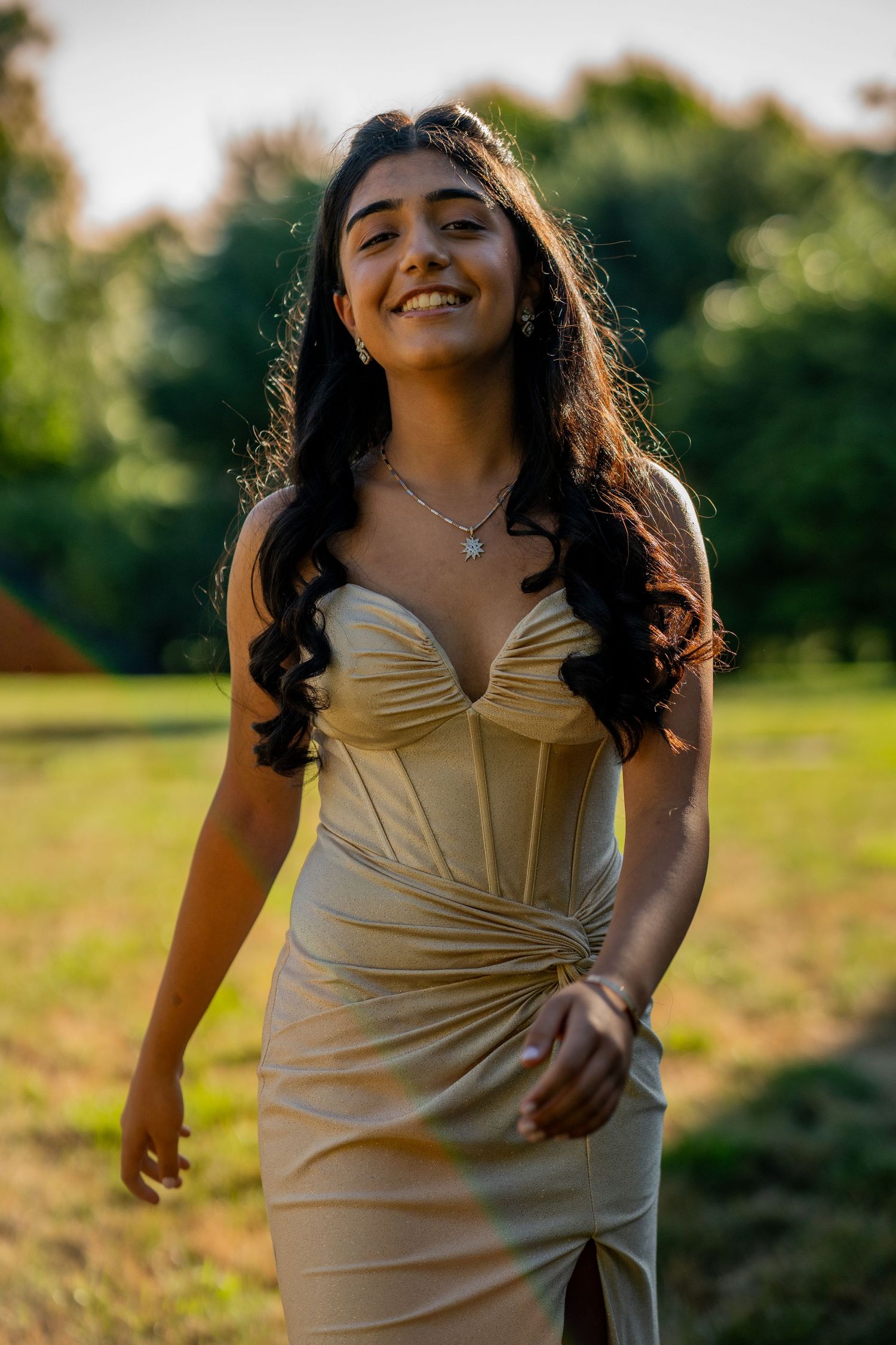 Young woman in gold strapless dress smiling outdoors at sunset with lush green trees in background.