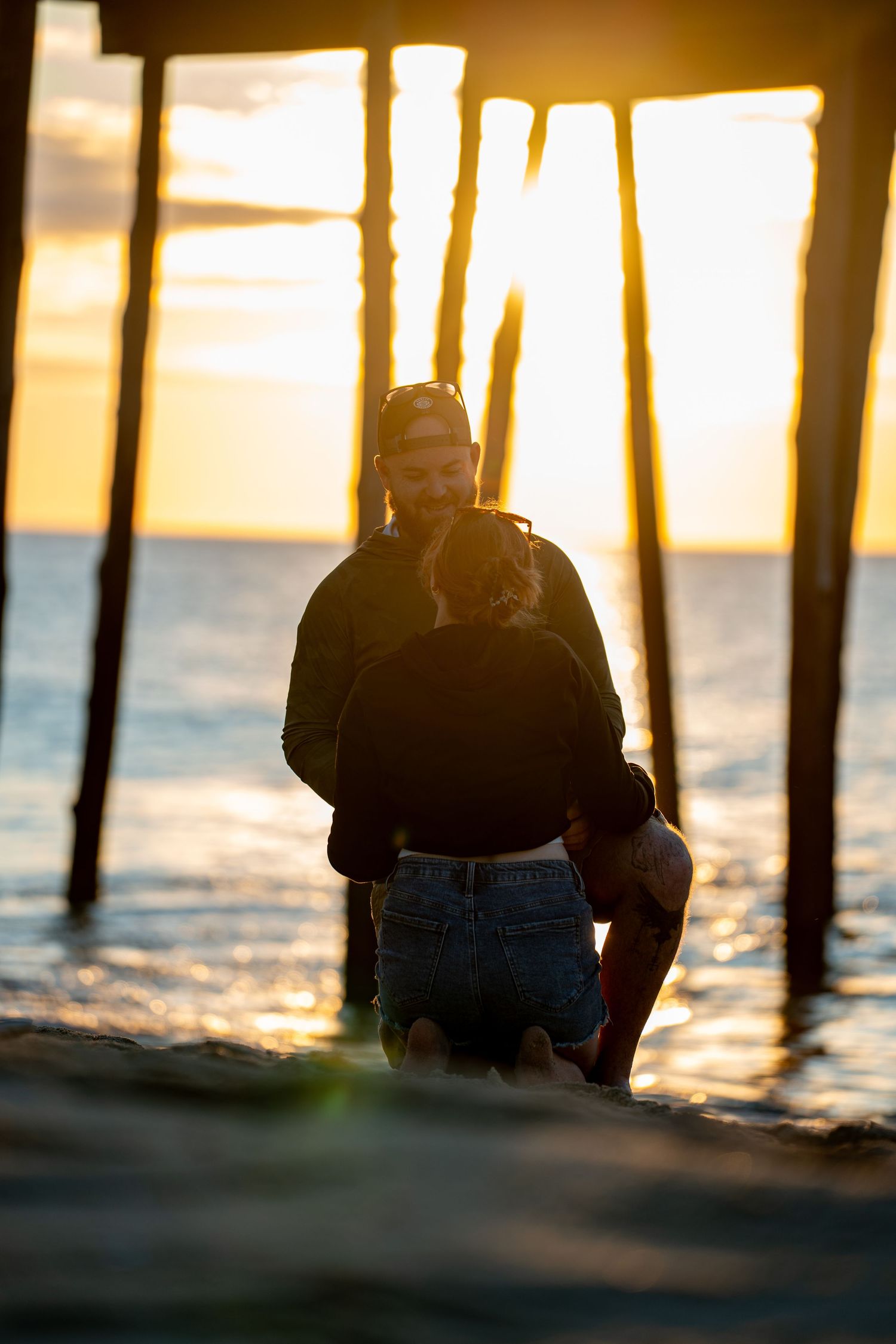 Silhouetted couple embracing on the beach beneath a wooden pier as the golden sunset glows over the calm ocean waves.