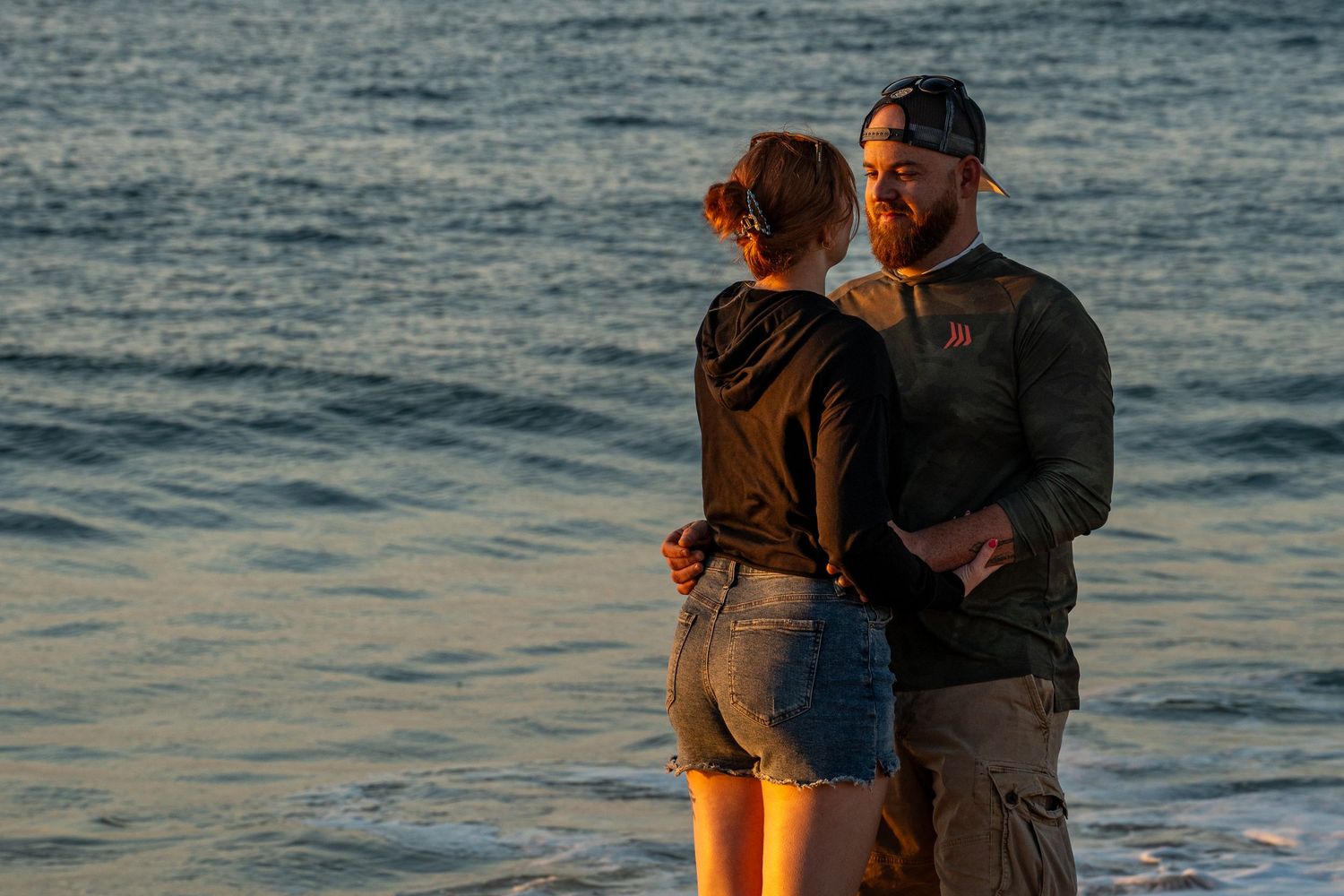 Couple embracing on the beach at sunset, the man in a cap and beard holding a woman in denim shorts as ocean waves roll in behind them.