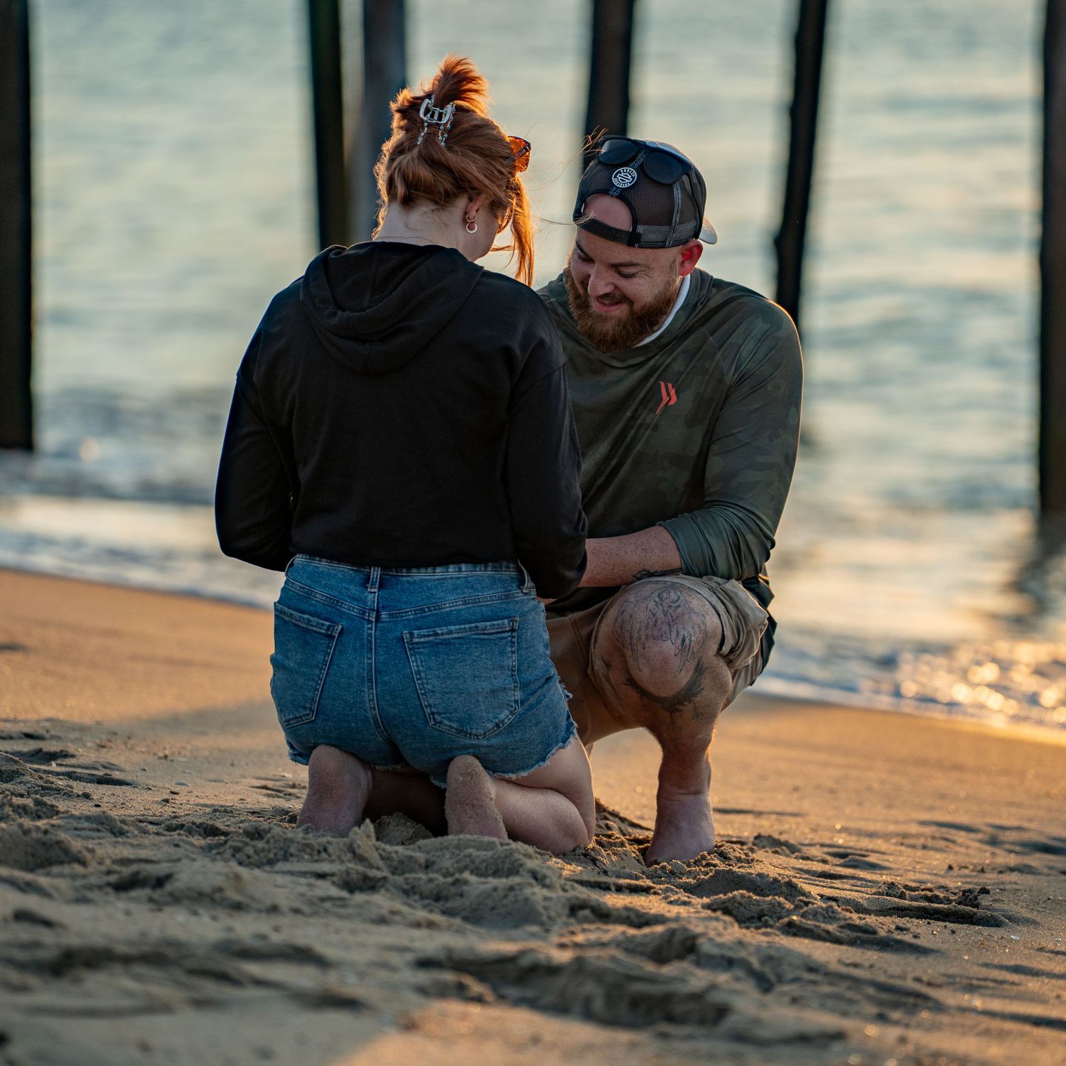 Couple kneeling face to face on a sandy beach at sunset beside wooden pier pilings, with calm ocean water glowing in the background.