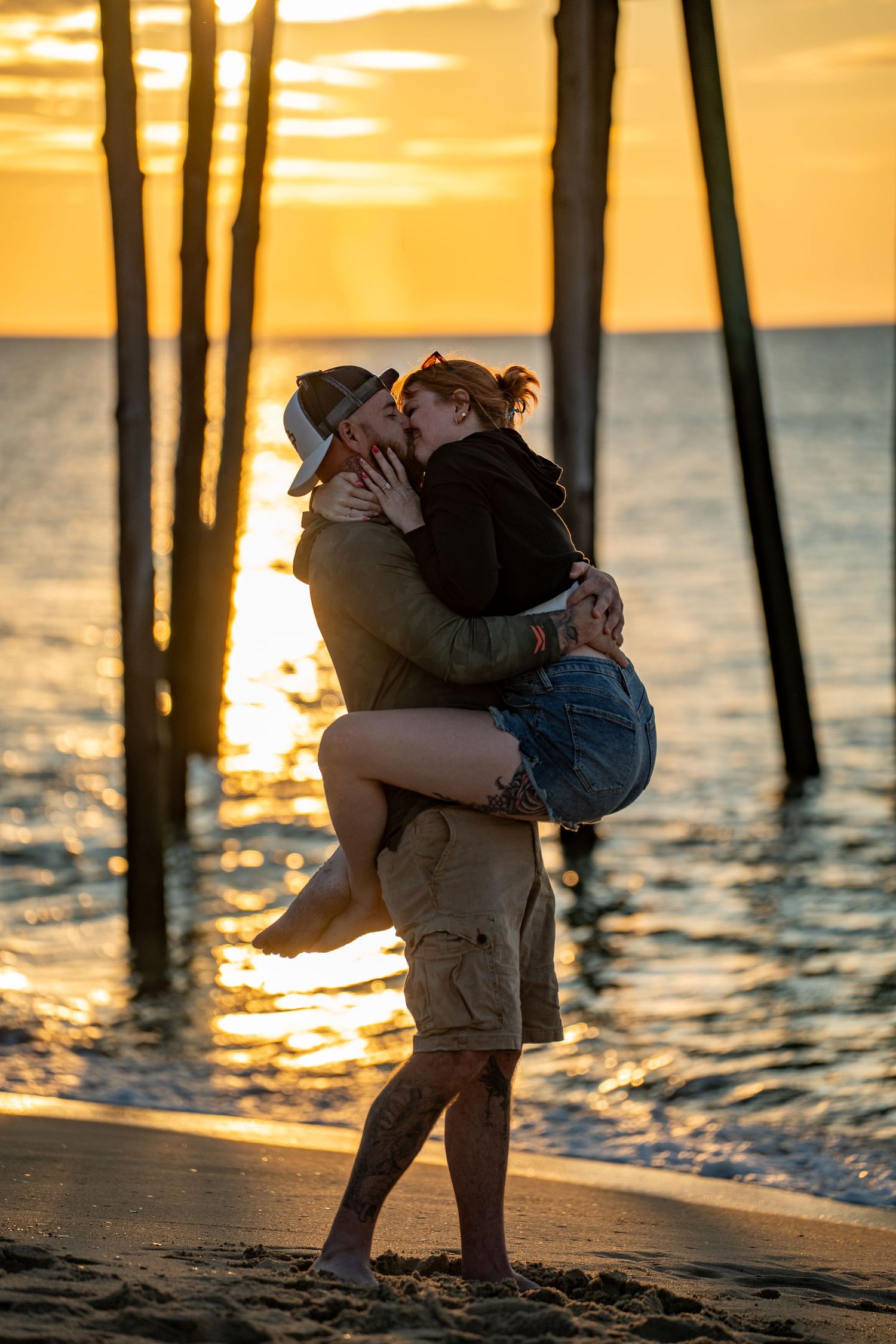 Couple kissing on the beach at sunset as he lifts her in his arms, with wooden pier posts and golden light reflecting on the ocean.