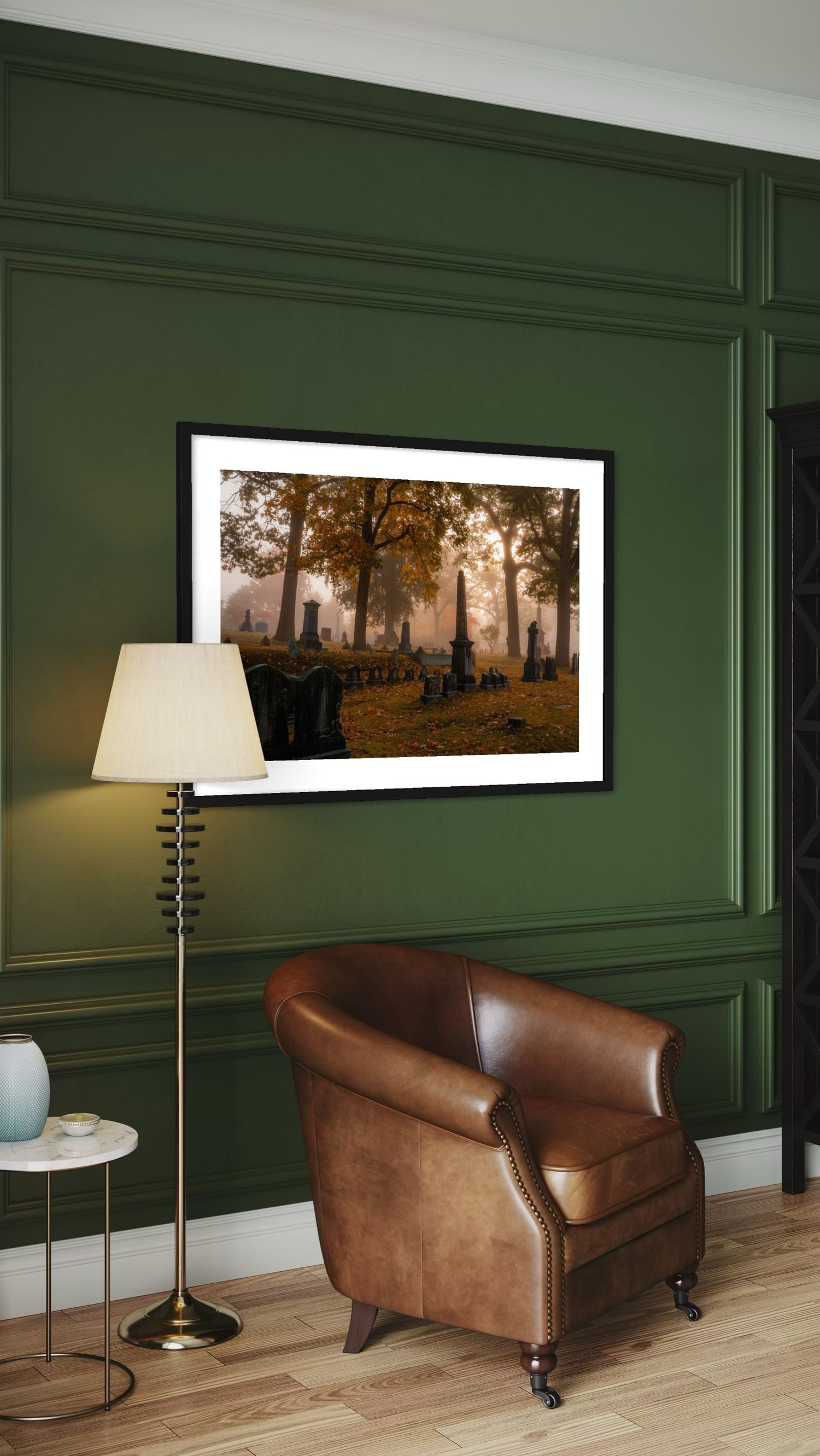 Framed autumn cemetery photo on green paneled wall above brown leather armchair with brass floor lamp.