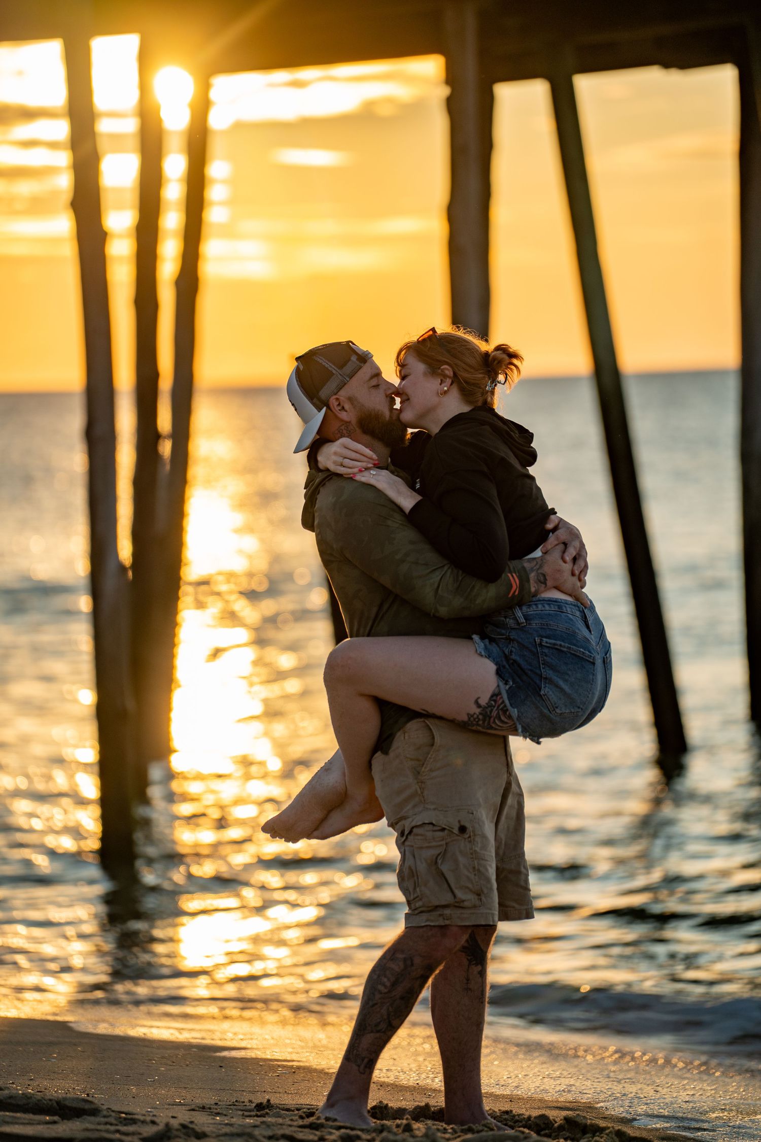 Couple embracing and kissing on the beach at sunset beneath a wooden pier, with golden light shimmering on the ocean waves.