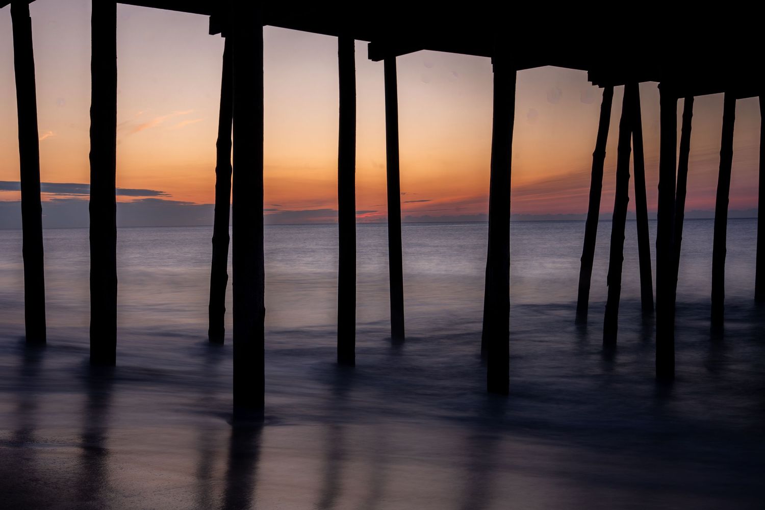 Silhouetted wooden pier pilings stand over calm ocean water at sunset, with soft orange and purple skies reflecting on the waves.