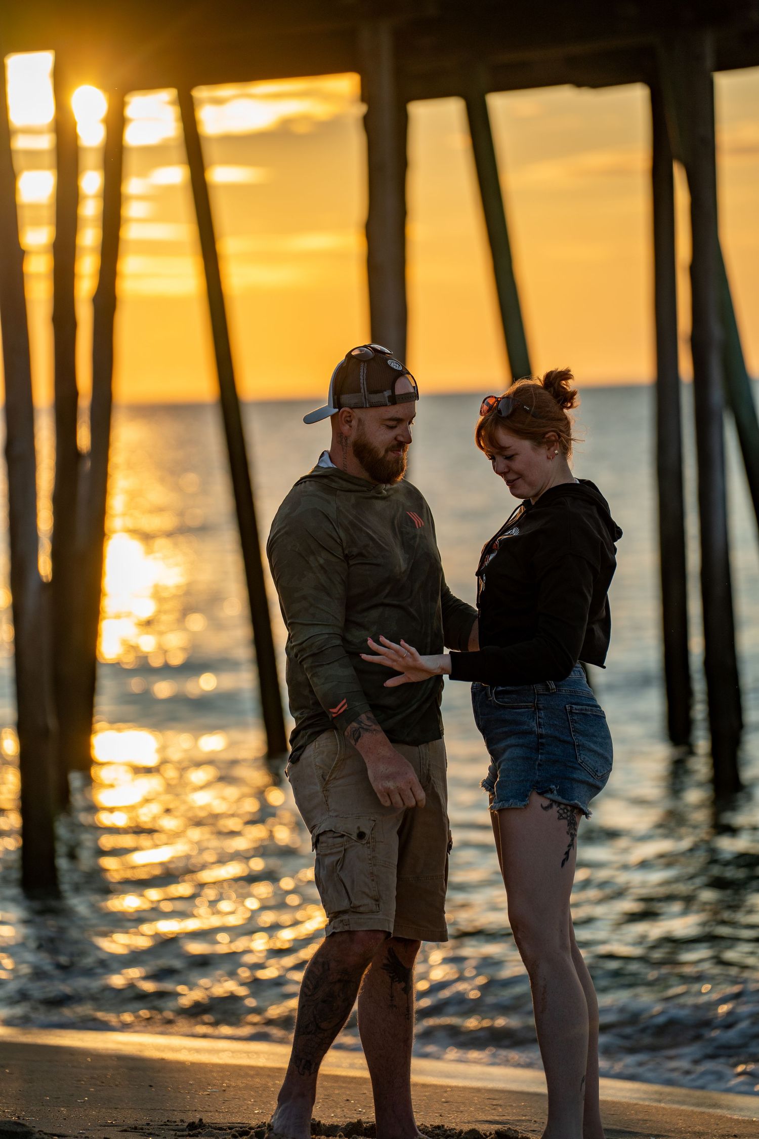 Couple standing close on the beach at sunset beneath a wooden pier, holding hands as golden light sparkles on the ocean waves.