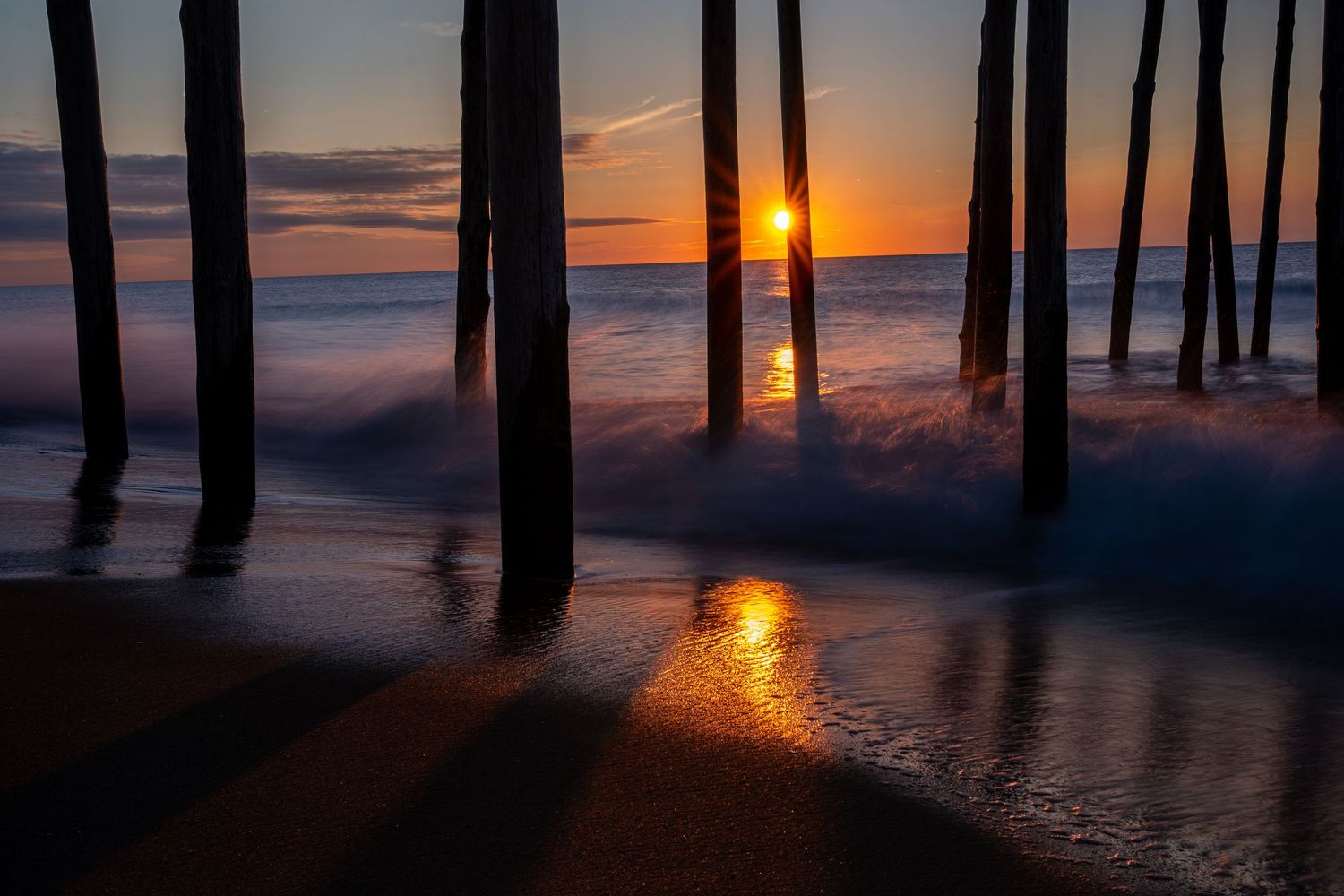 Stunning sunset viewed under a pier with waves washing ashore, casting golden reflections on wet sand between wooden pilings.