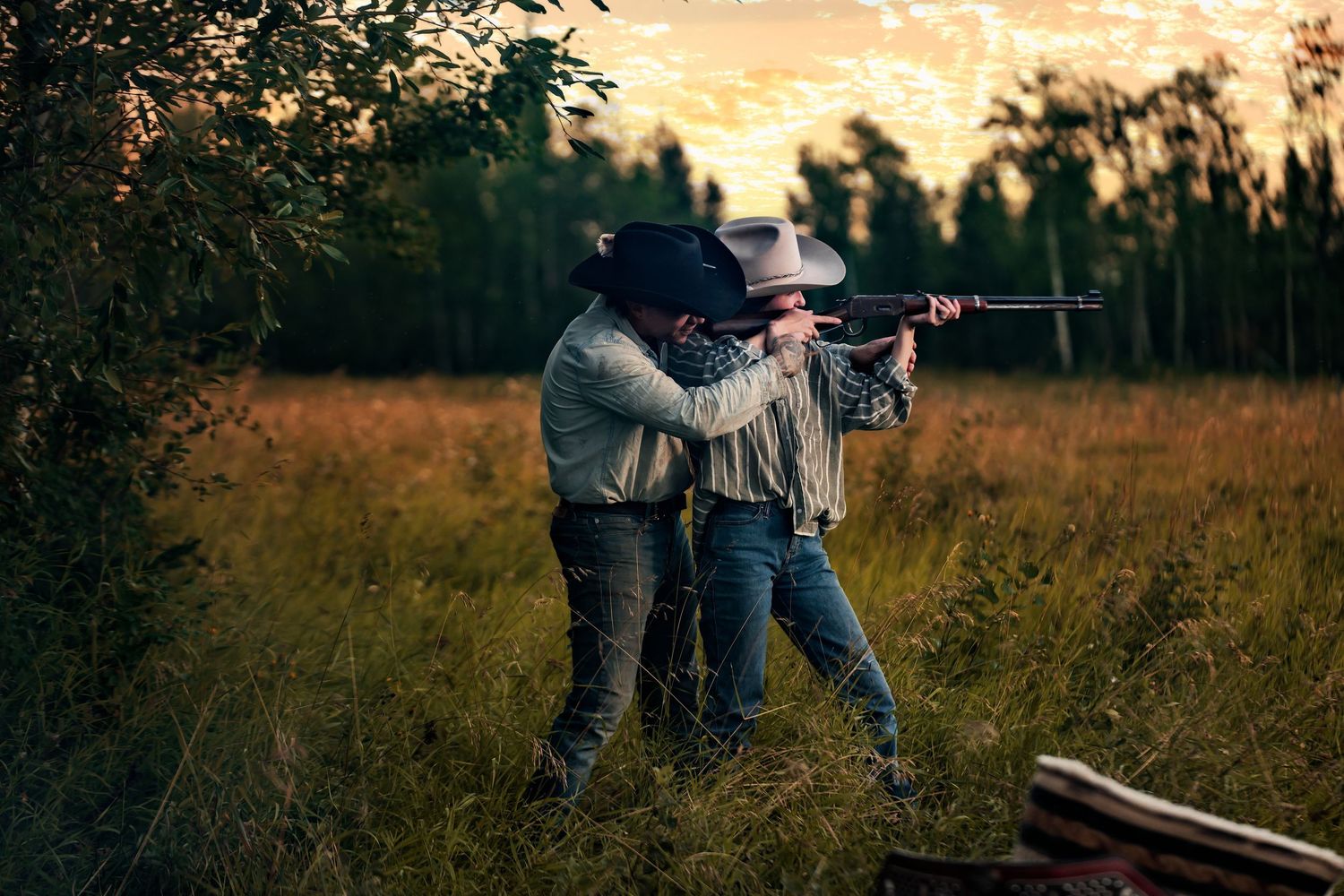 Two cowboys aiming a rifle in a golden field at dusk, surrounded by trees and tall grass.