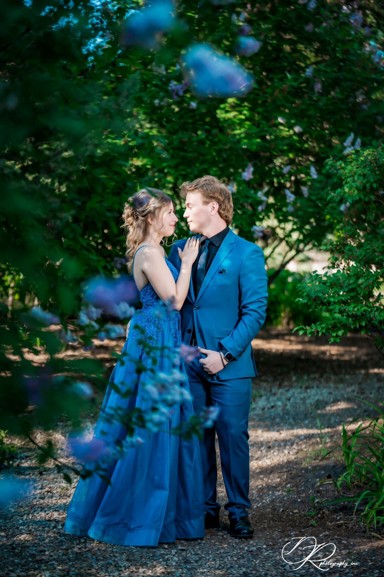 A couple in matching blue formal wear share a romantic moment surrounded by lush greenery and purple blooms.