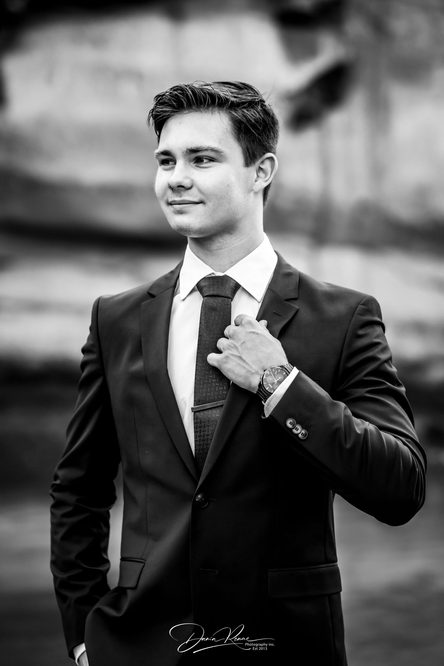Black and white portrait of a young man in a suit adjusting his tie, wearing a watch, smiling confidently outdoors.