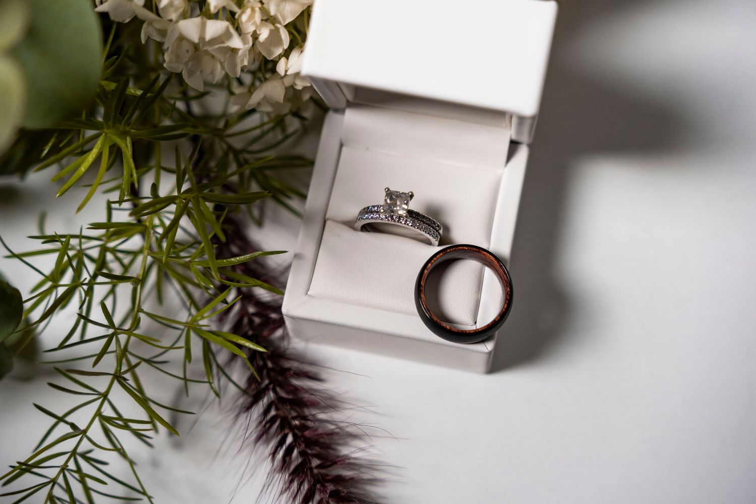 Wedding rings displayed in a white box with diamond engagement ring and wooden band beside green floral arrangement.