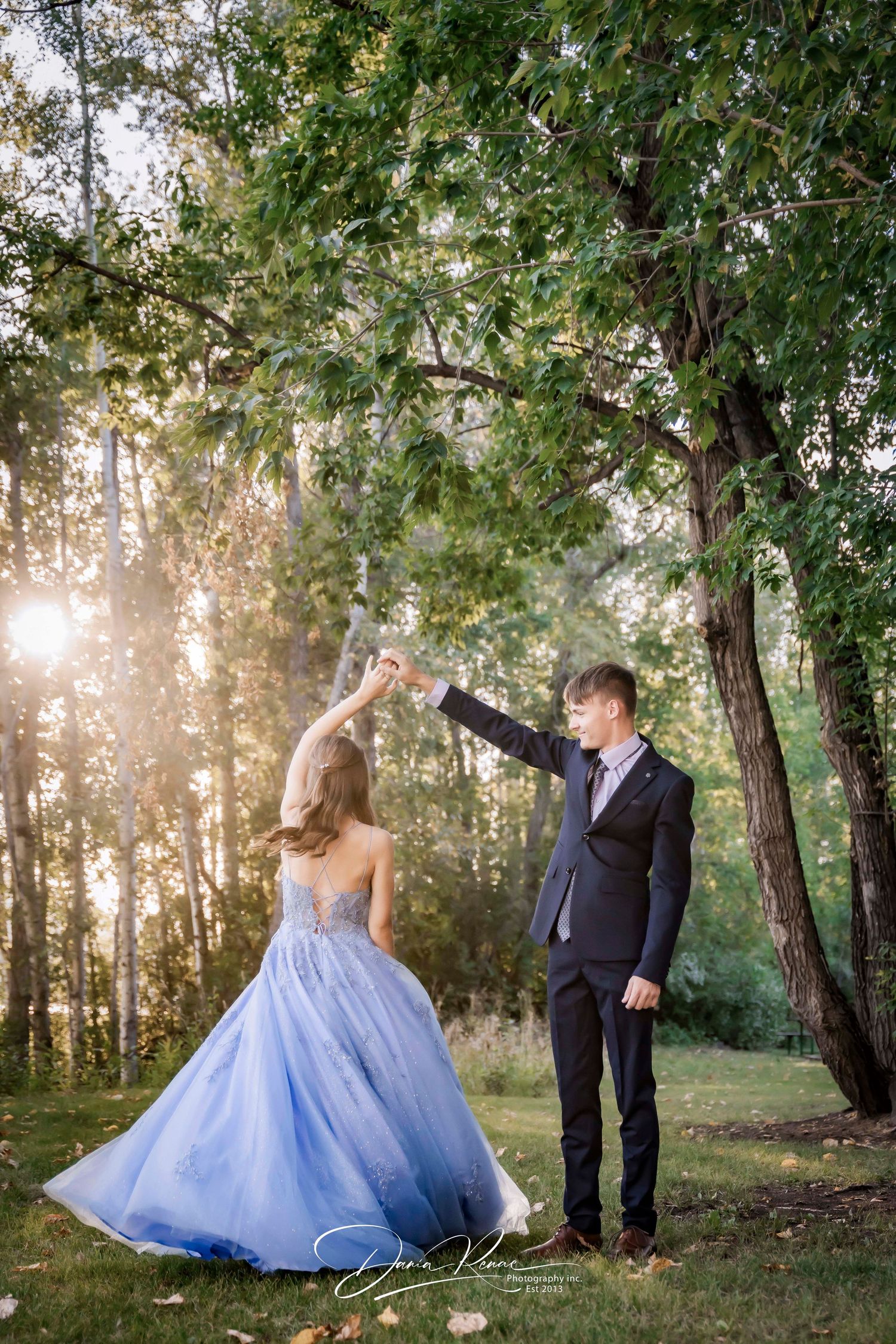 A couple dances outdoors in golden sunlight, woman in blue ball gown, man in black suit, surrounded by lush green trees.