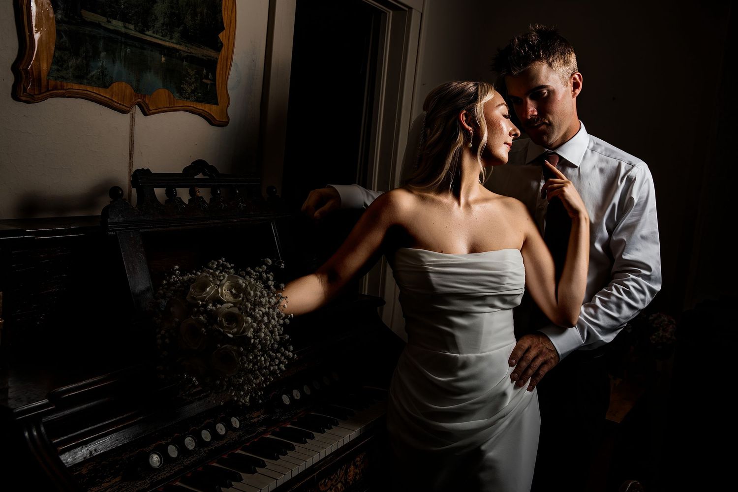 Romantic couple in elegant wedding attire sharing an intimate moment near a vintage piano in dramatic moody lighting.