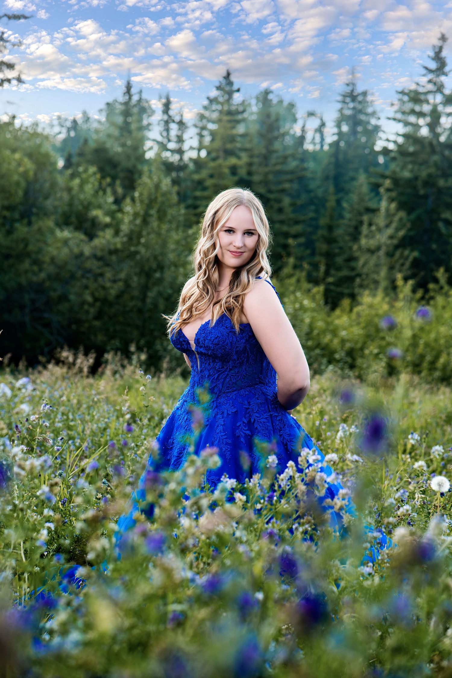 Young woman in a royal blue gown poses in a wildflower meadow with evergreen trees and a cloudy sky.