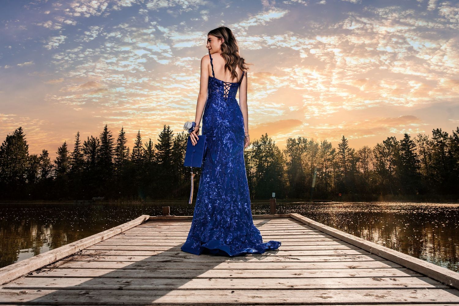 Woman in royal blue sequin gown standing on wooden dock at sunset with forest and lake backdrop.