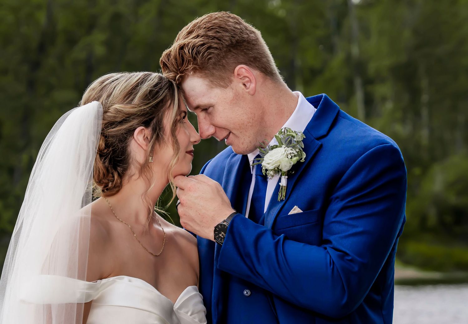 A groom in a blue suit tenderly kisses his bride's hand as they share an intimate moment outdoors on their wedding day.