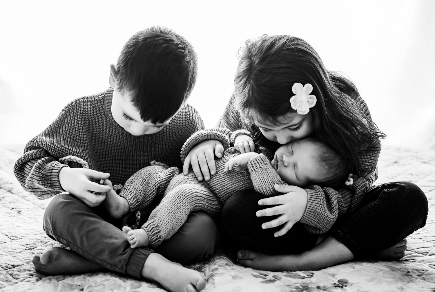 Black and white photo of two older siblings lovingly holding and kissing a sleeping newborn baby.