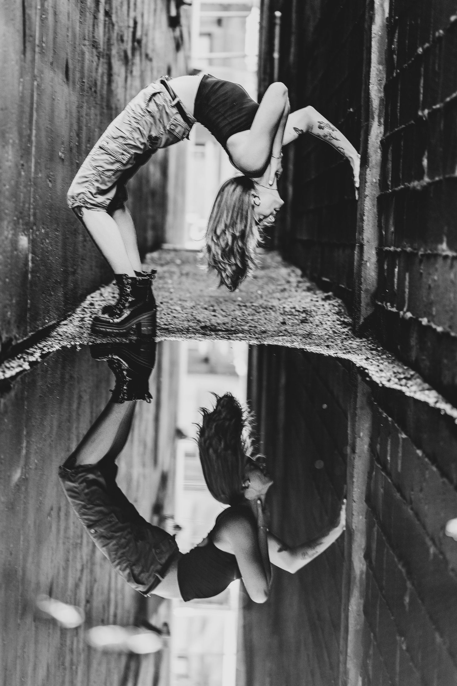 Black and white photo of a woman doing a backbend in an alley, reflected in a puddle below.