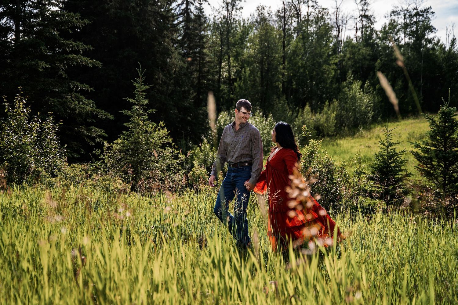 Couple holding hands in a lush green meadow surrounded by evergreen trees on a sunny day.