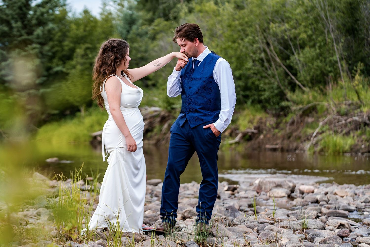 Groom in blue vest kisses bride's hand outdoors near rocky riverbank surrounded by lush green trees.