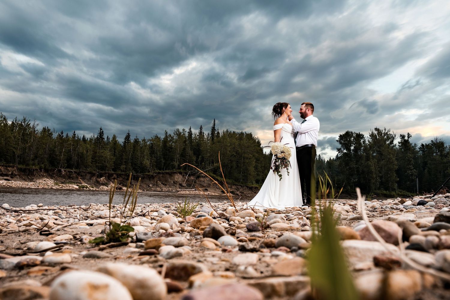 Bride and groom embrace on rocky riverbank with dramatic stormy sky and forest backdrop.