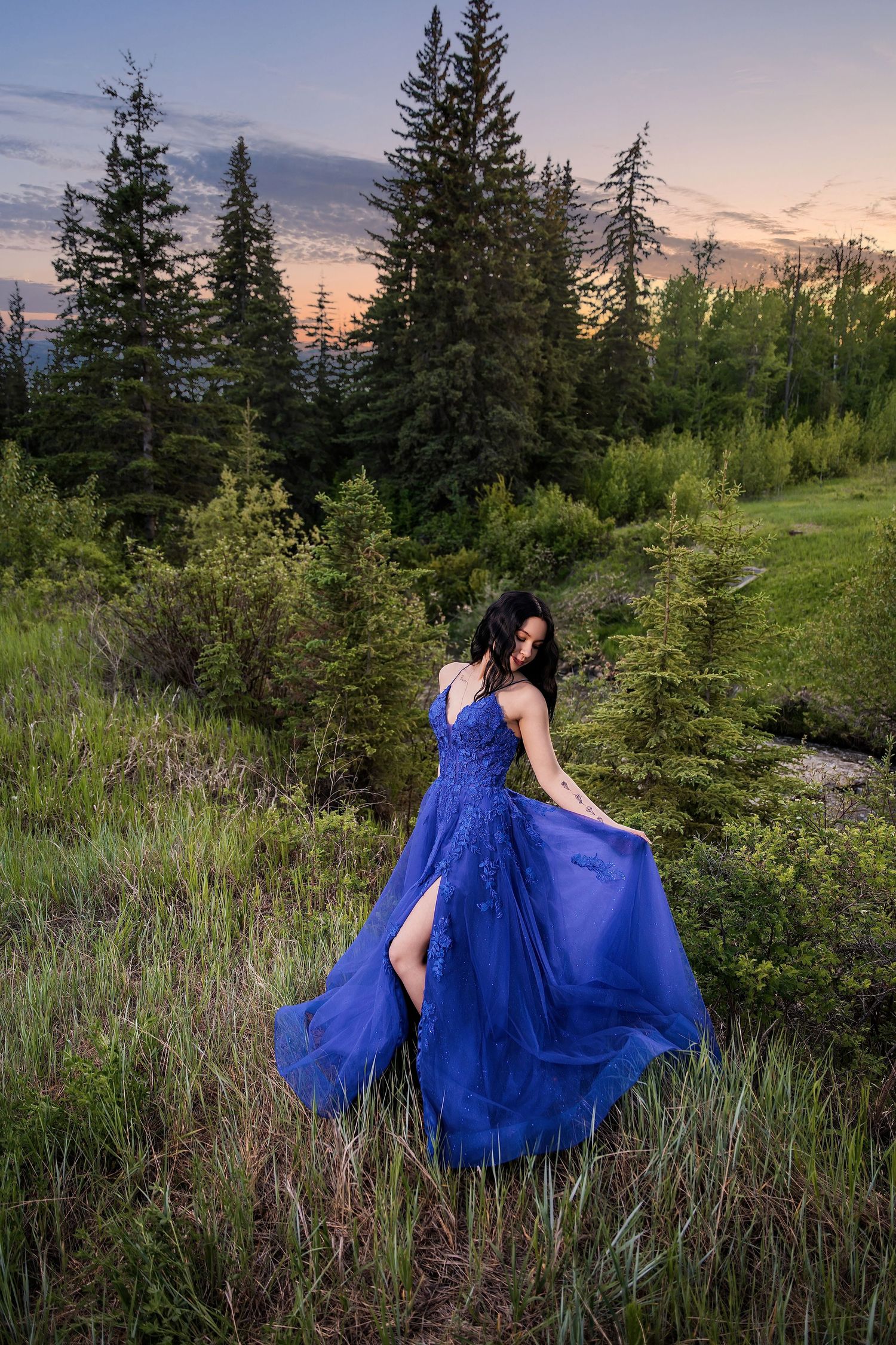 Woman in flowing blue gown poses gracefully in lush forest meadow at sunset.