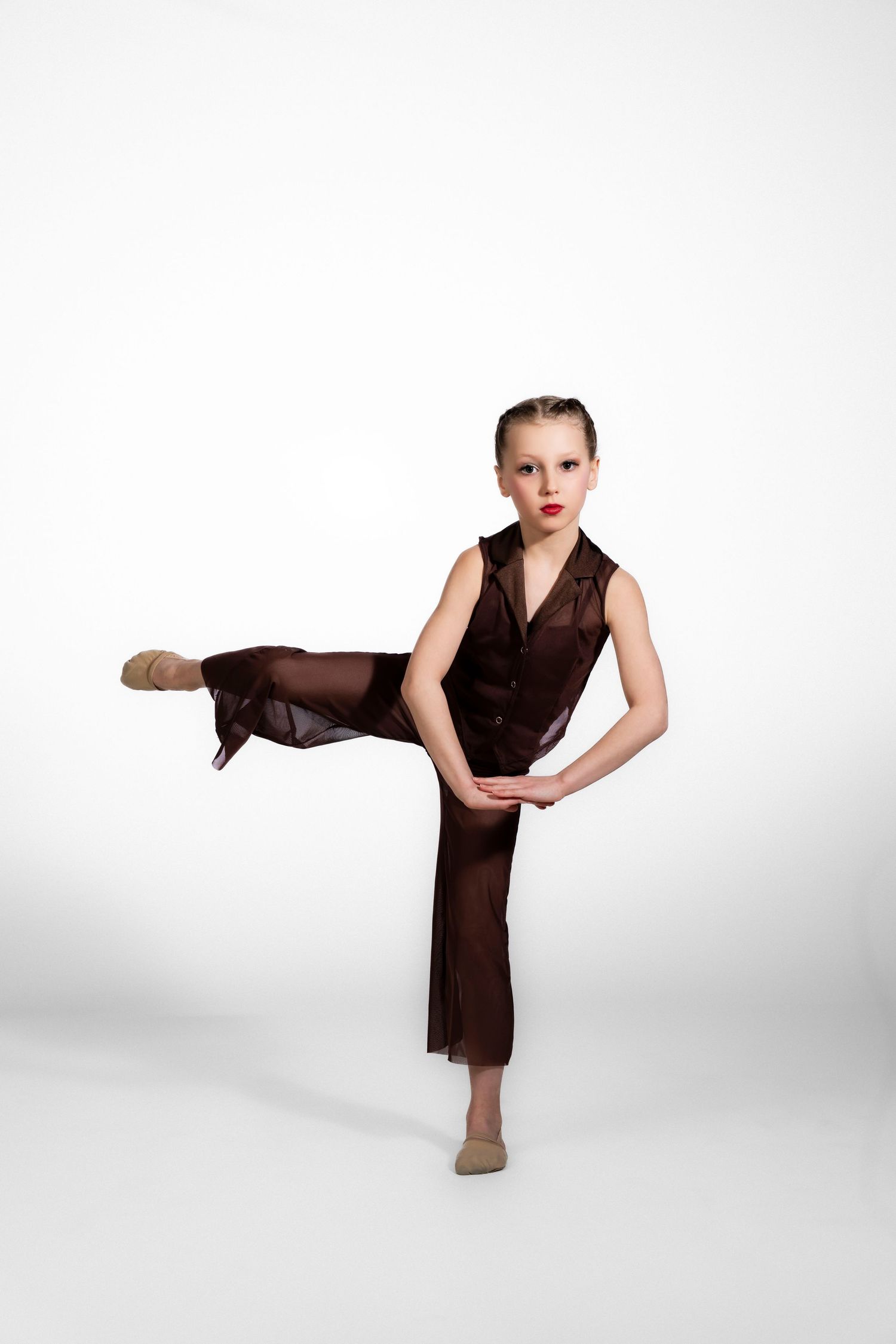 Young girl dancer in brown costume performs a high kick pose against white background.