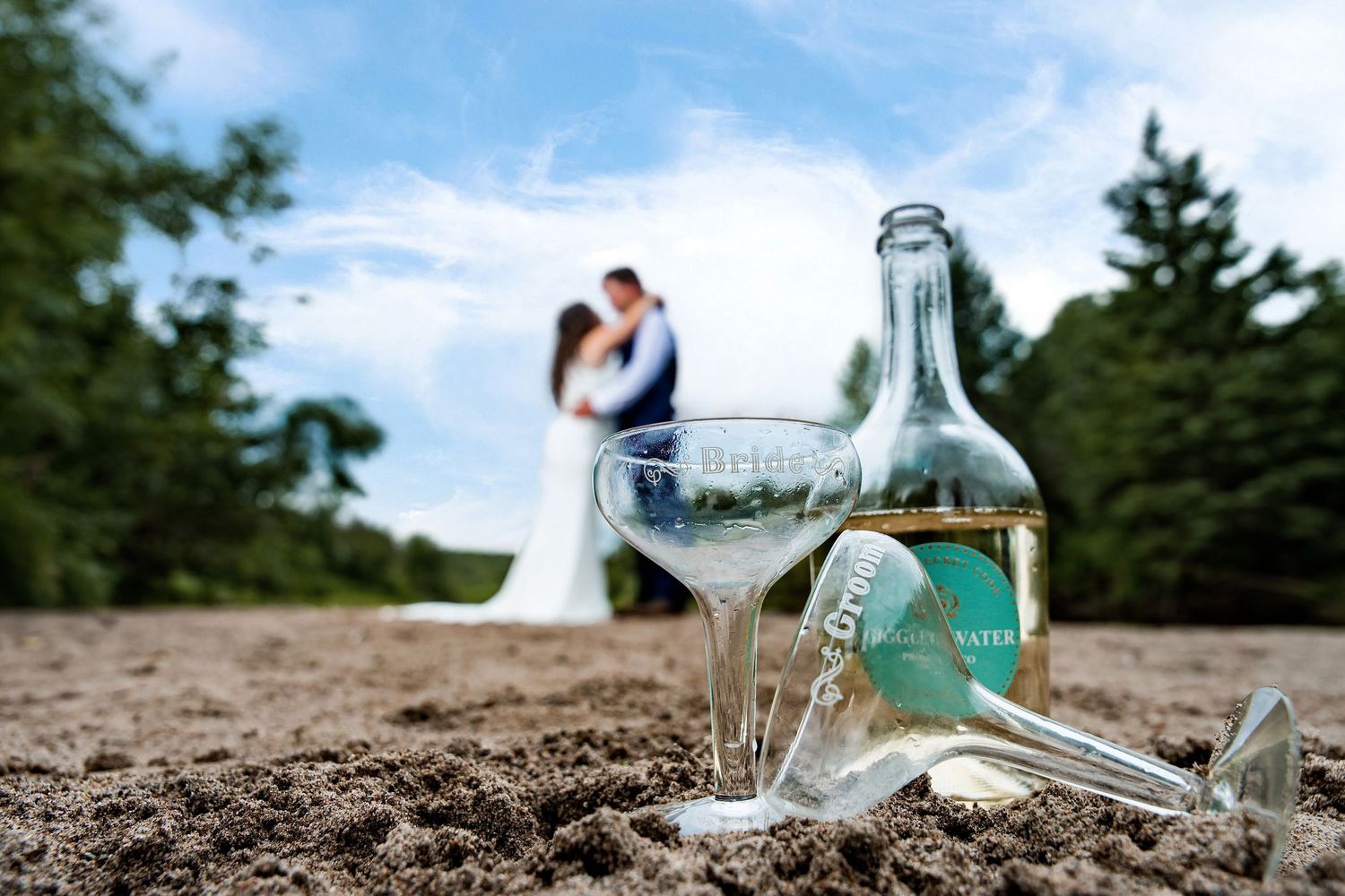 Wedding couple kissing on sandy beach with champagne bottle and martini glasses in foreground under blue sky.