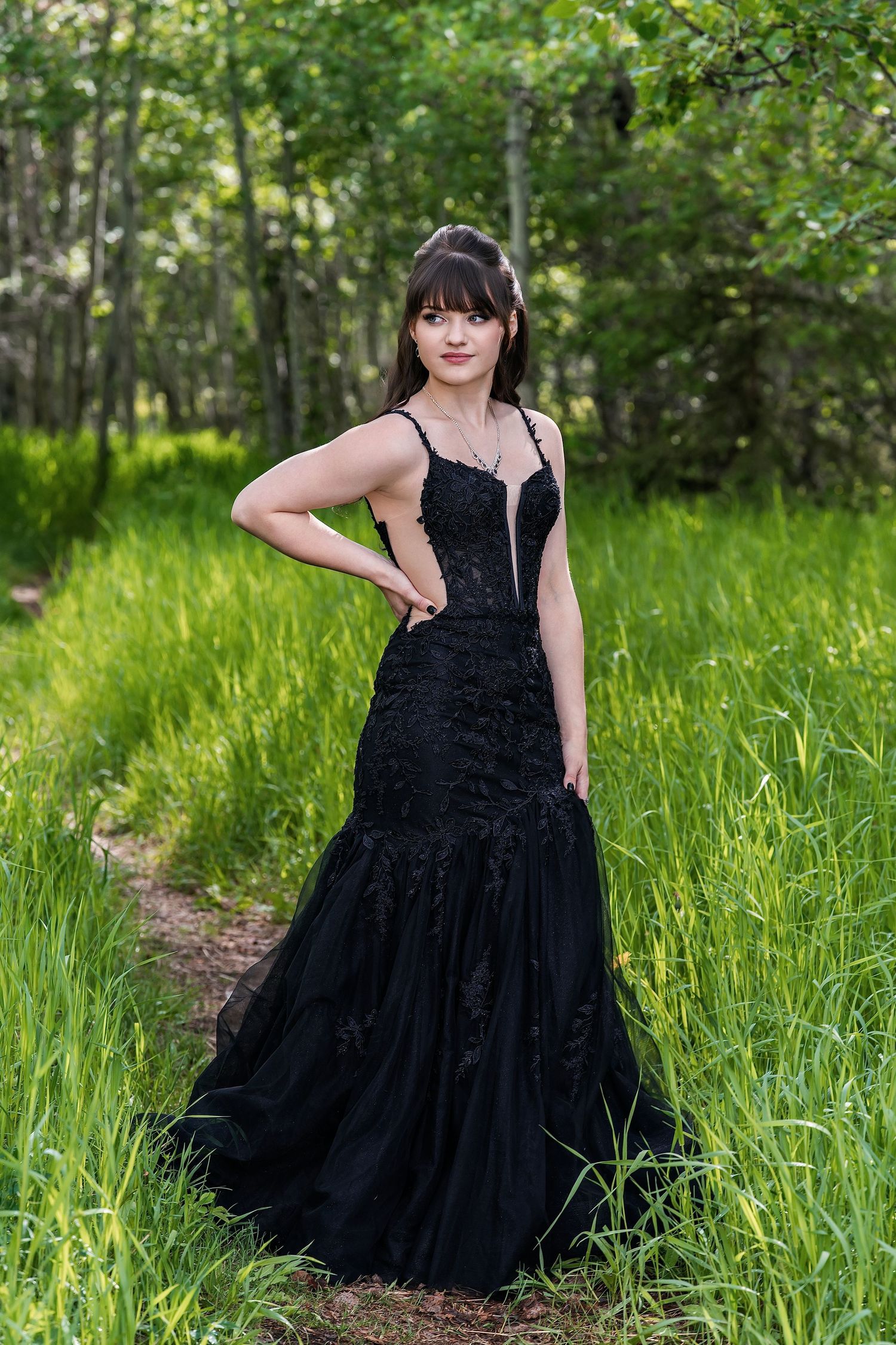 Young woman in elegant black mermaid gown poses gracefully on a forest path surrounded by lush green trees.