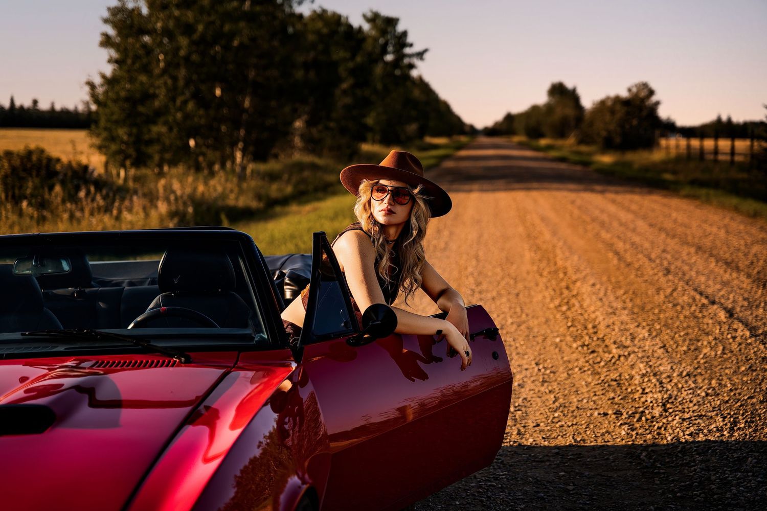Woman in cowboy hat and sunglasses leaning on red convertible sports car on rural dirt road at sunset.