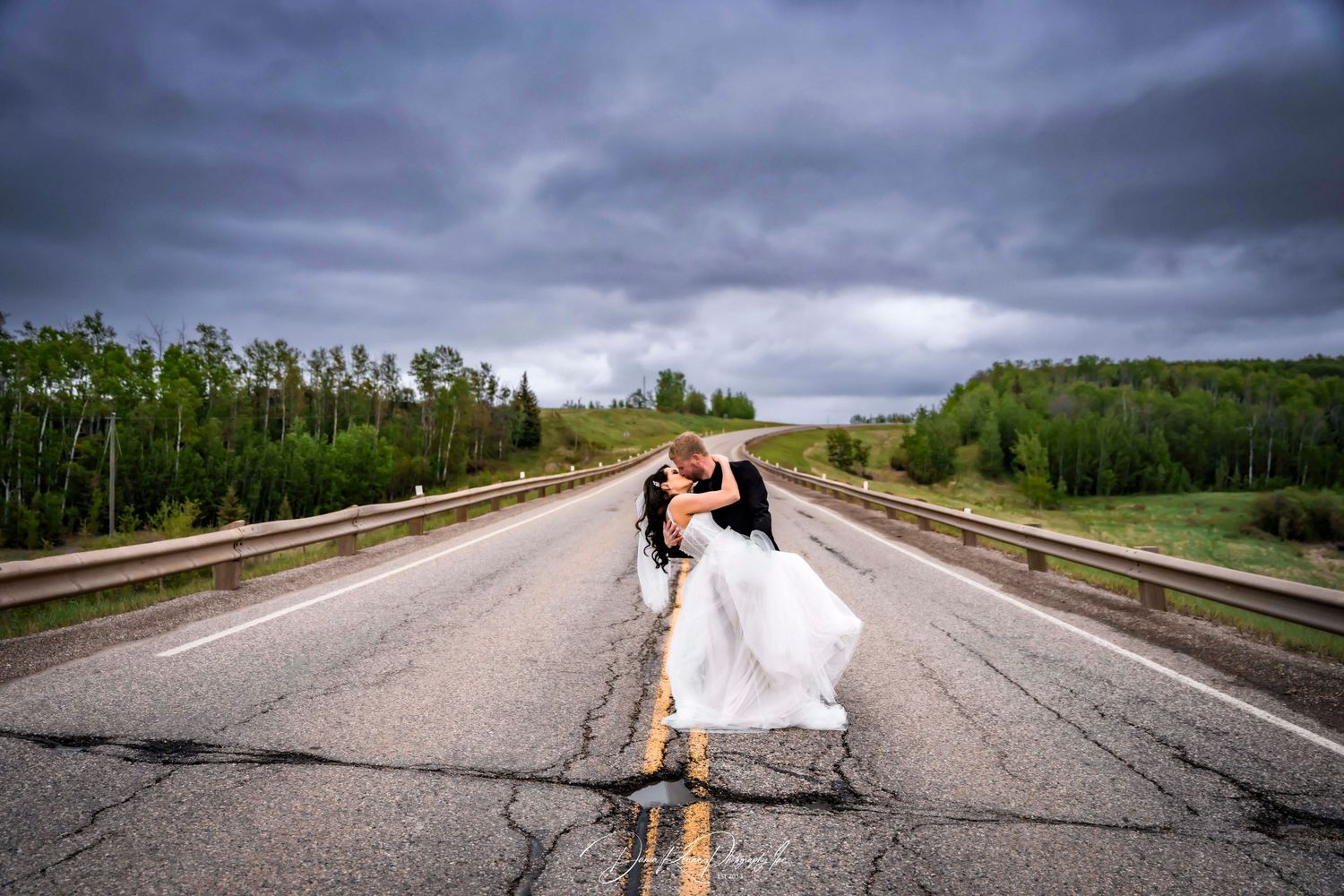 Bride and groom share a romantic dip kiss on an empty highway under dramatic stormy skies surrounded by lush green trees.