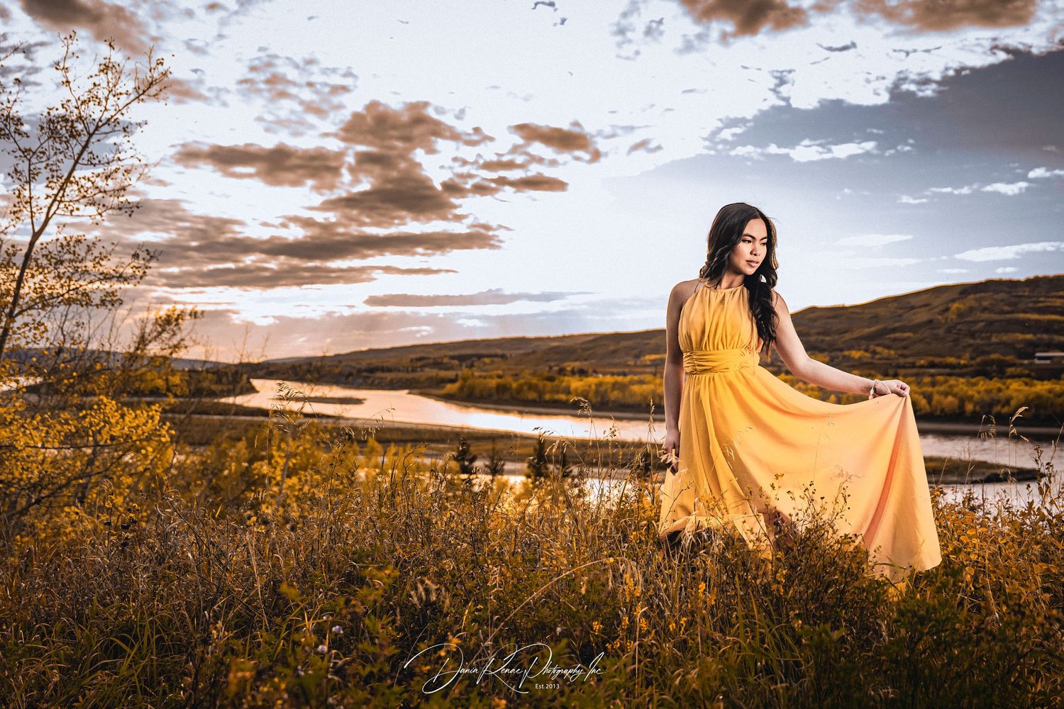 Woman in yellow dress posing in golden field at sunset with river and dramatic cloudy sky backdrop.