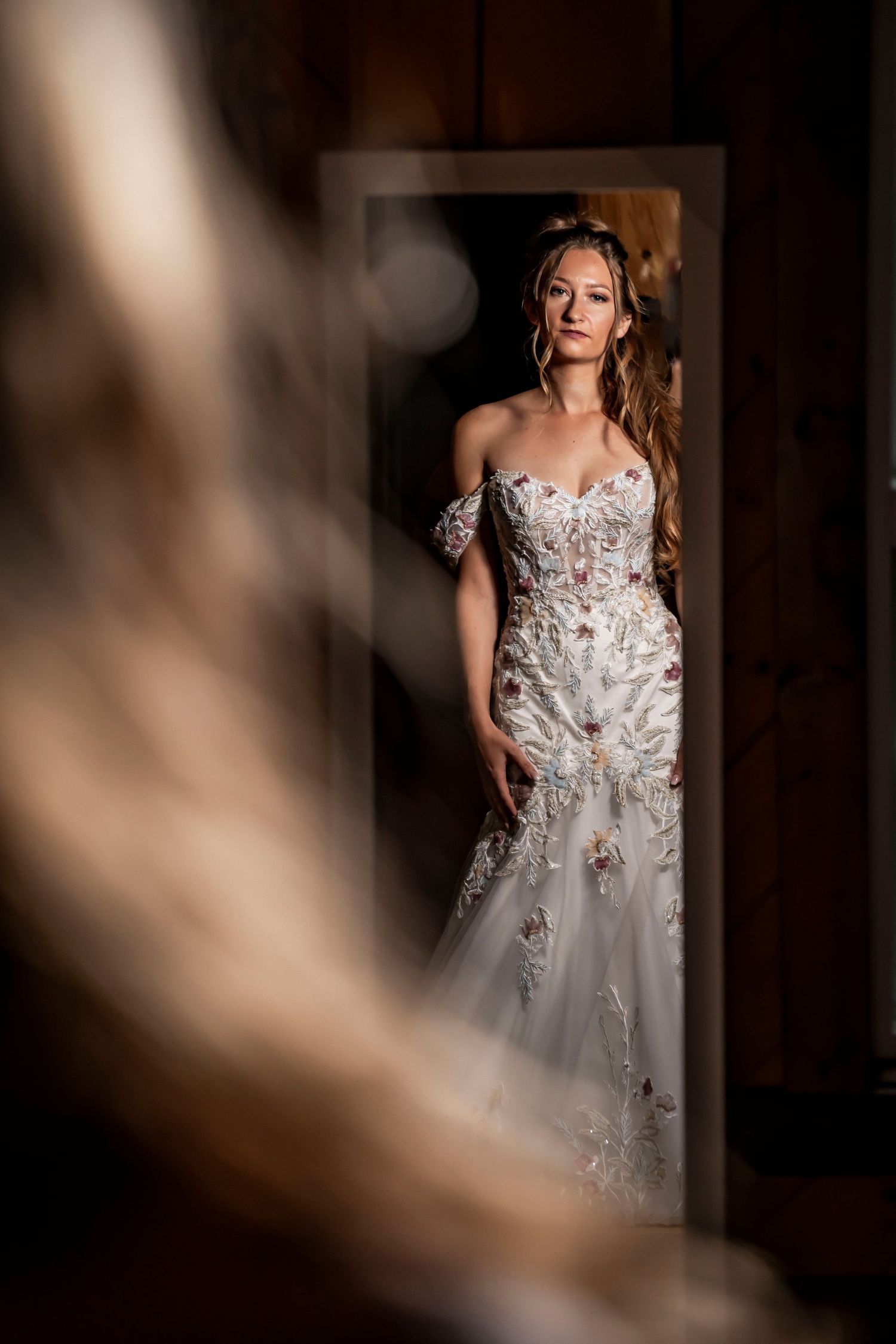 Bride in floral off-shoulder wedding gown stands elegantly in a doorway, captured in a dramatic mirror reflection.