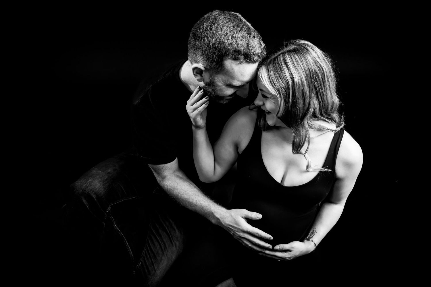 Black and white maternity photo of couple embracing tenderly, woman cradling baby bump in elegant studio portrait.