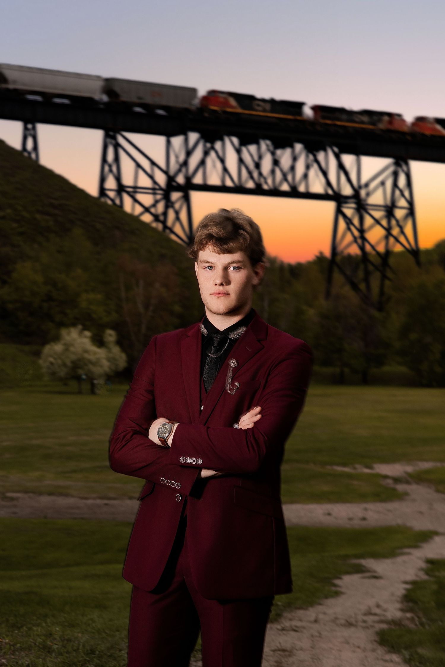 Young man in burgundy suit stands arms crossed before a railroad trestle bridge at sunset.