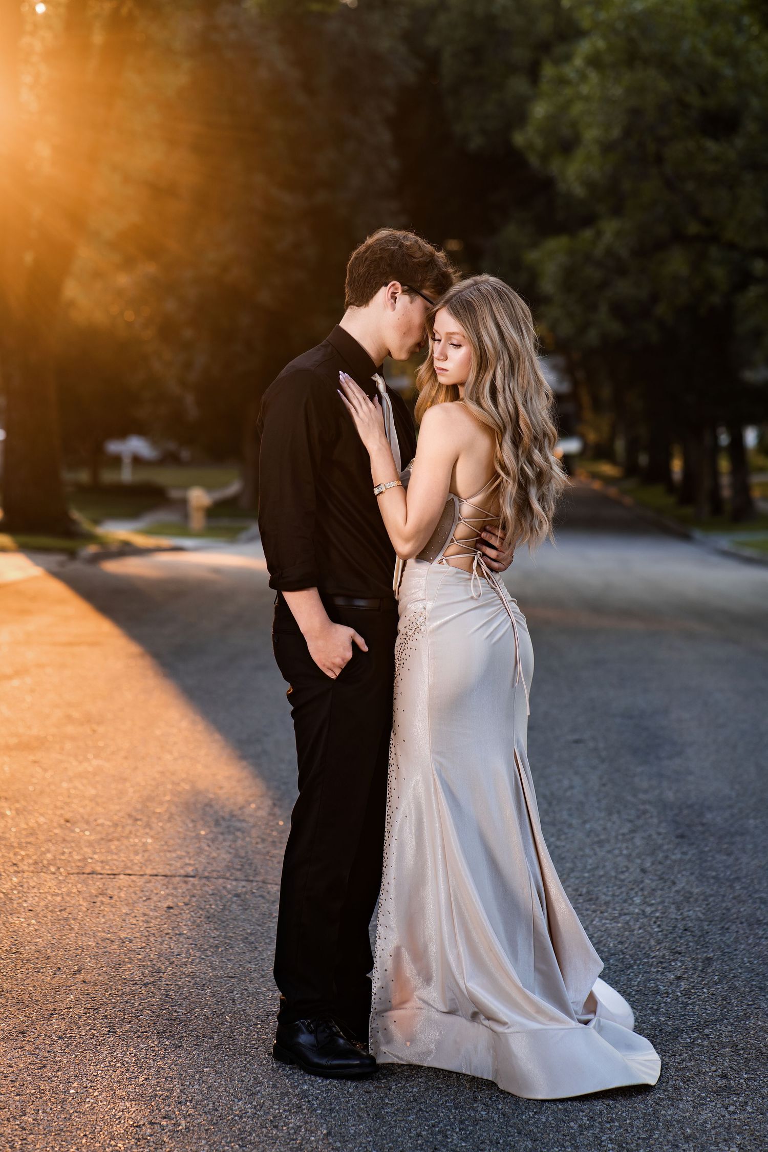 Couple in formal attire embracing on a sunlit road at golden hour, woman in elegant backless gown, man in black suit.