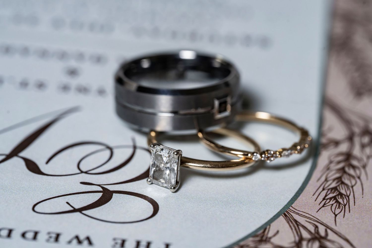 Gold diamond engagement ring, wedding band, and silver groom ring displayed on elegant wedding invitation.